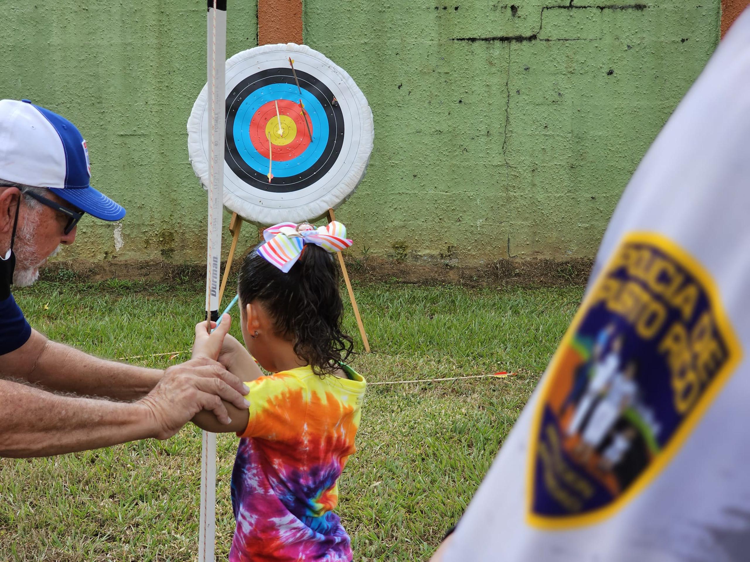 Una niña aprende el deporte de arco y flecha, mientras un agente de la Policía observa como parte del impacto comunitario que realizan en el residencial Pedro Zorrilla de Manatí.