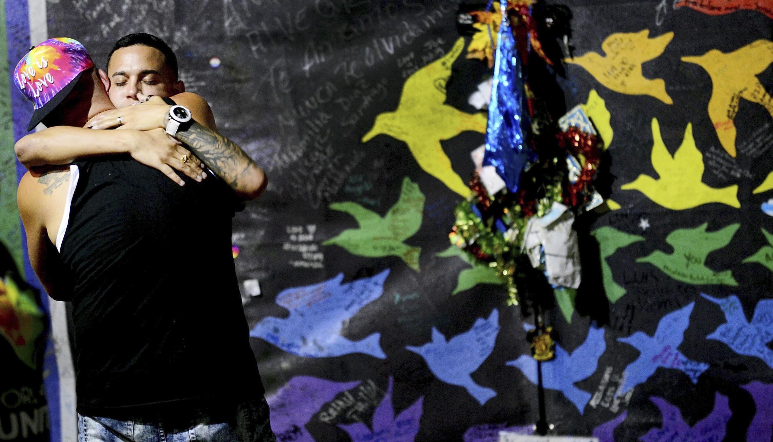 Dos participantes de los actos conmemorativos se abrazan, dándose apoyo emocional, frente al mural que recoge los rostros de las víctimas. (EFE / Gerardo Mora)