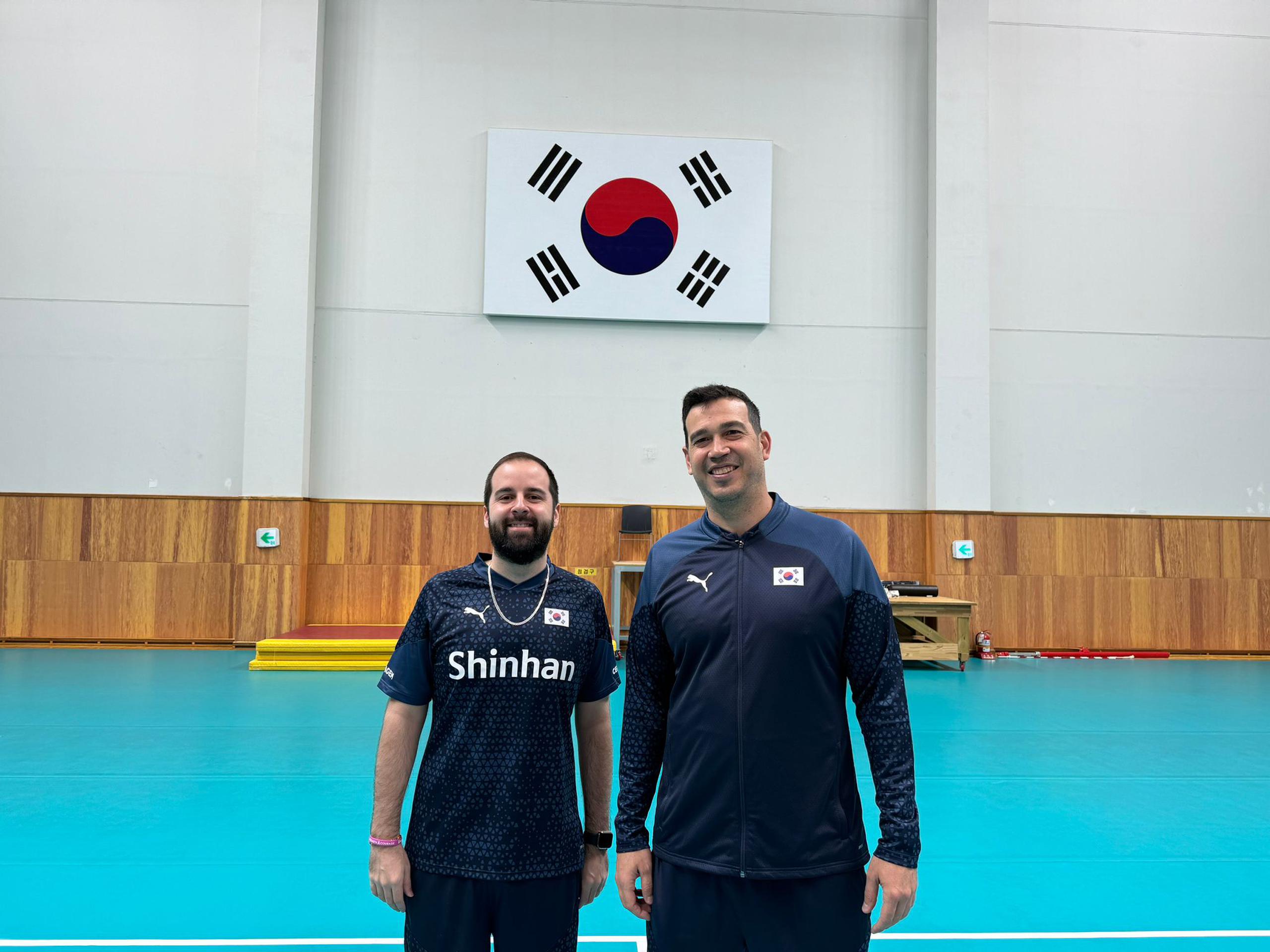 Fernando Morales y Jesús Echevarría a la derecha e izquierda, respetivamente, posan en la cancha de entrenamientos en Corea del Sur con la bandera de Corea del Sur al fondo.