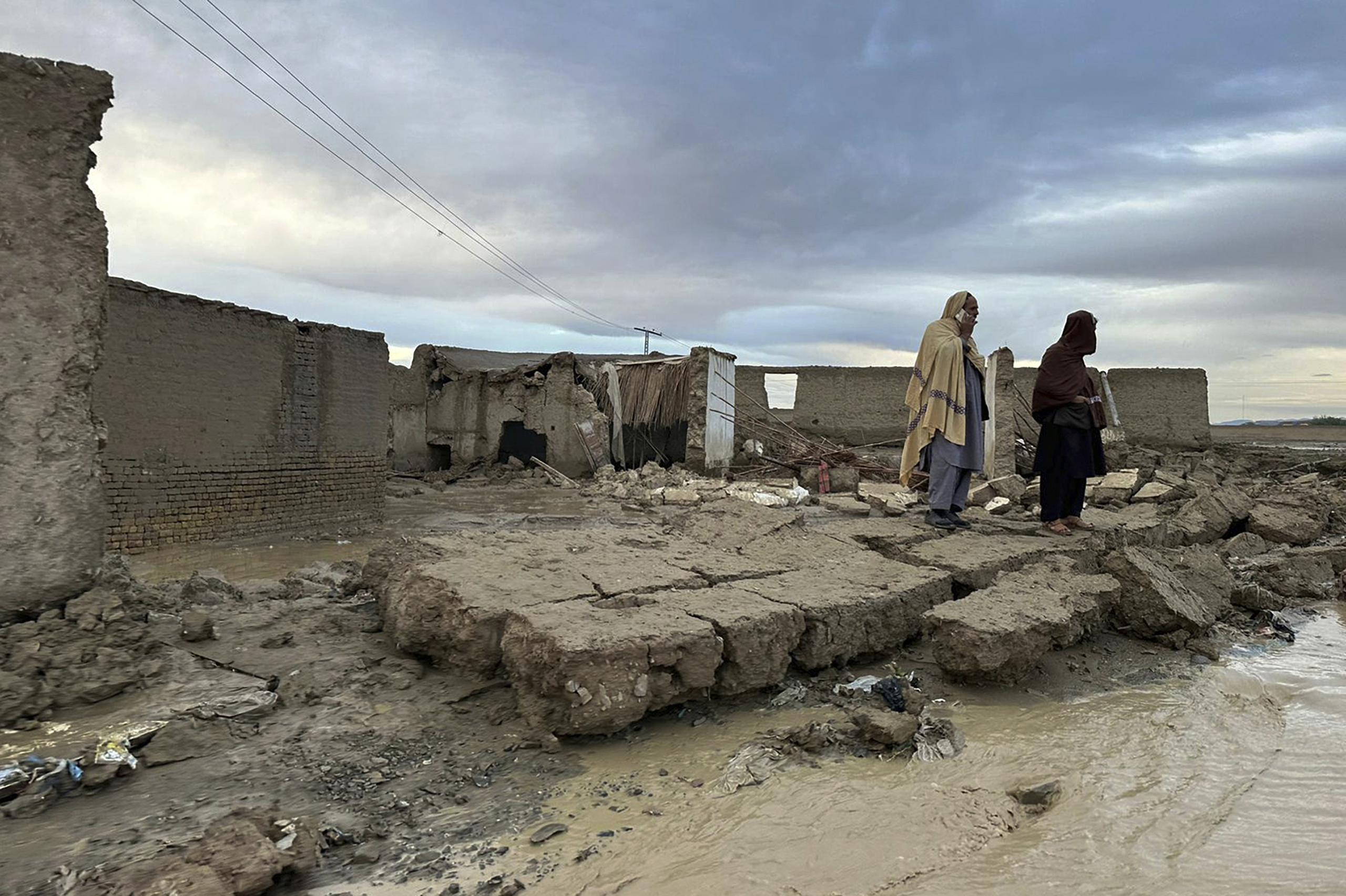 Personas permanecen de pie sobre un muro caído de su vivienda, que sufrió daños debido a las fuertes lluvias en un área cerca de Chaman, Pakistán, el jueves, 18 de abril de 2024. (AP Foto/Habib Ullah)