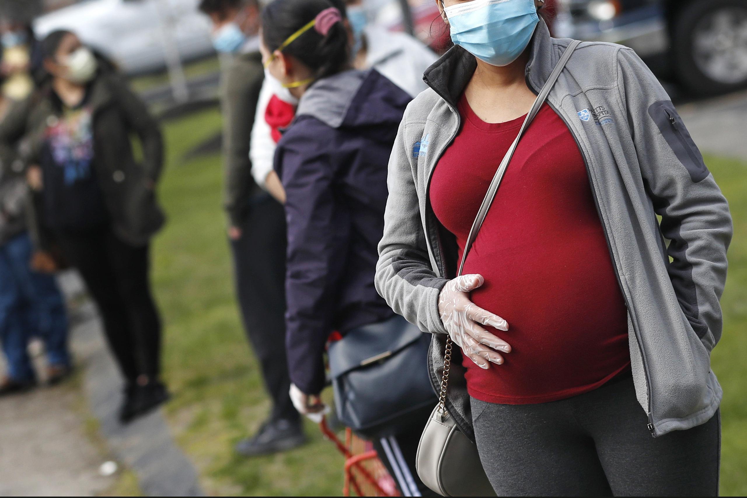Una mujer embarazada que utiliza mascarilla y guantes por la pandemia de coronavirus hace fila con numerosas personas para recibir una despensa de alimentos financiada por la organización caritativa Healthy Waltham en la iglesia St. Mary en Waltham, Massachusetts