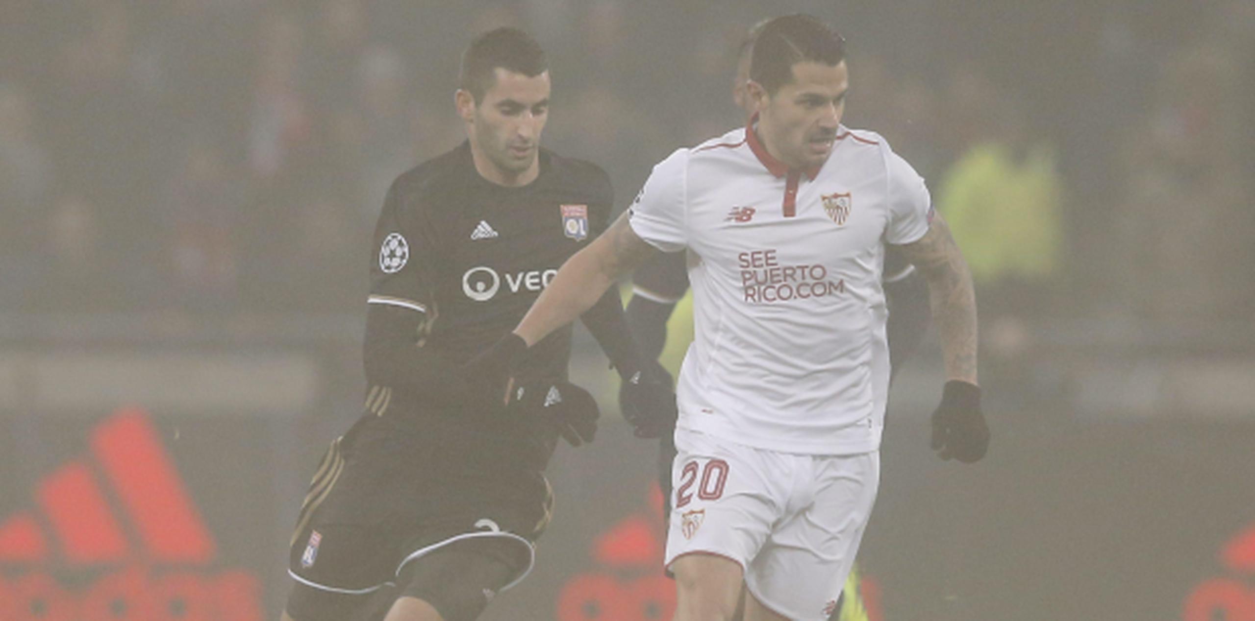 Maxime Gonalons del Olympique Lyonnais disputa el balón con Vitolo del Sevilla FC en el campo arropado por neblina. (Agencia EFE)