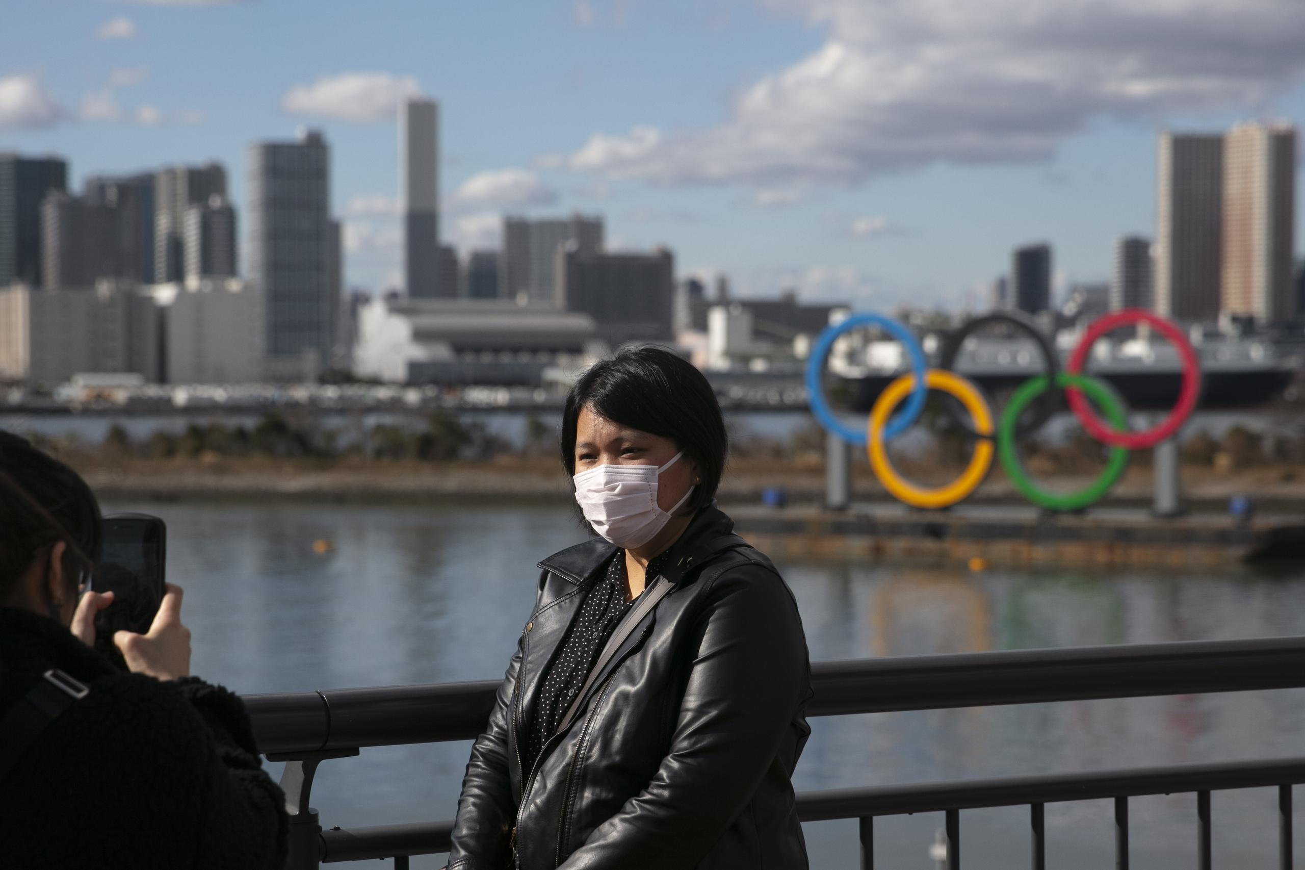 En esta foto una turista con un cubreboca posa para una foto, con los aros olímpicos al fondo, en el distrito de Odaiba, en Tokio.