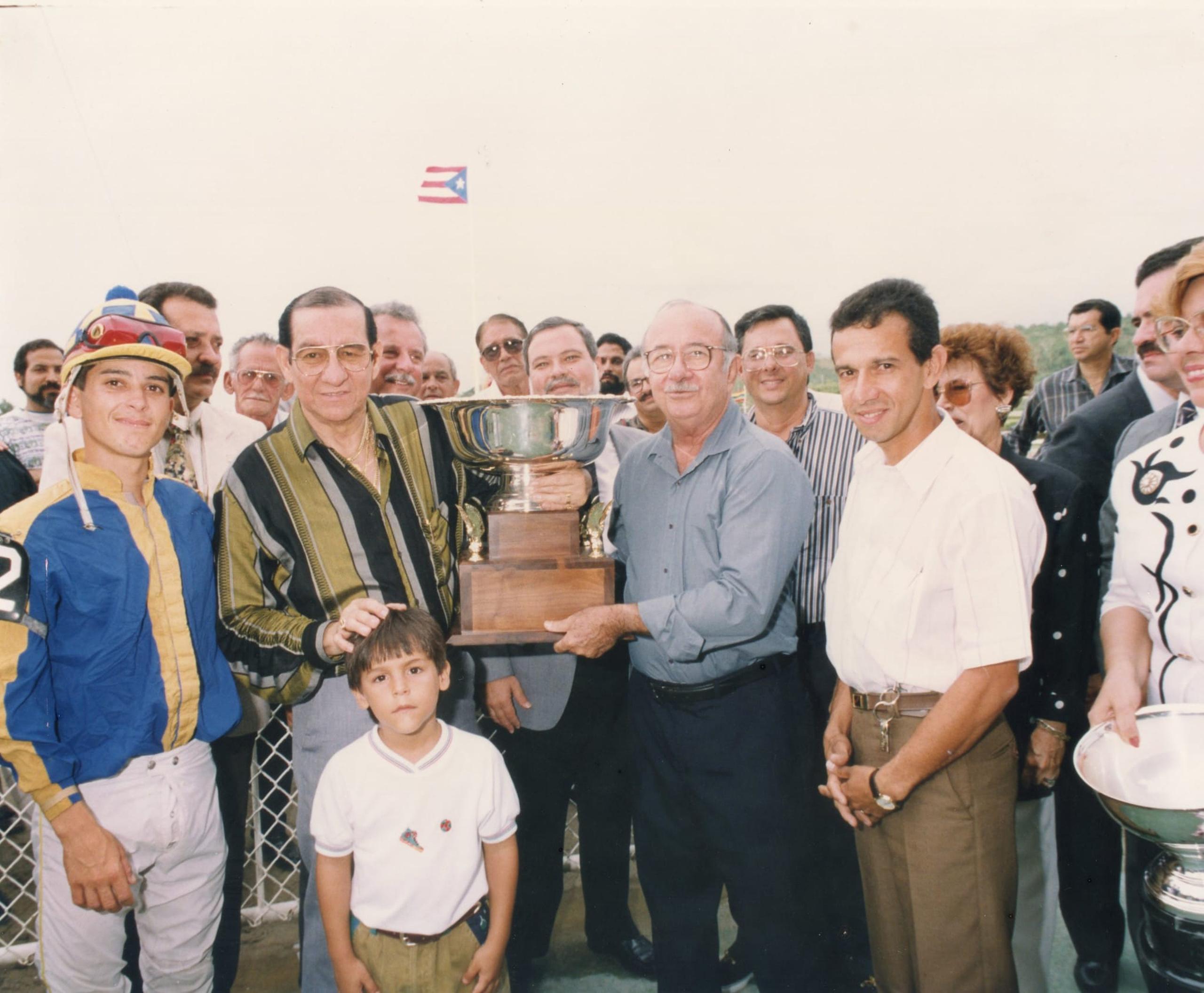 Tommy Muñiz, con camisa gris,  entrega la Copa San Juan a Luis Hernández por su triunfo en el Clásico San Juan del 1991 en el Hipódromo El Comandante. La carrera fue ganada por Vuelve Candy B, con la monta de Alexis Feliciano. En ese evento el caballo completó la conquista de la Triple Corona.