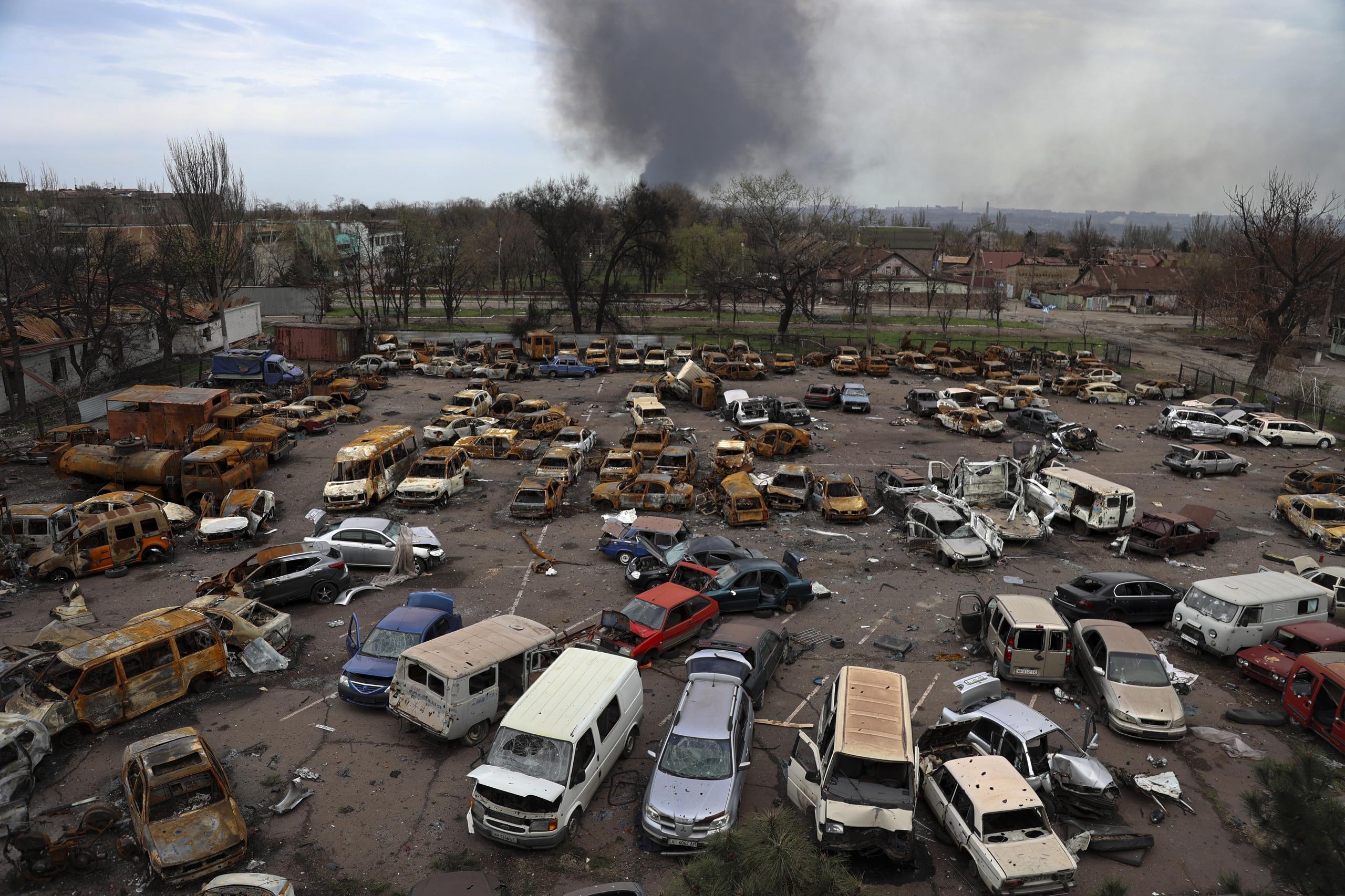 Autos destruidos en un estacionamiento próximo a la planta siderúrgica de Azovstal, en Maiupol, que se ve humeando al fondo.