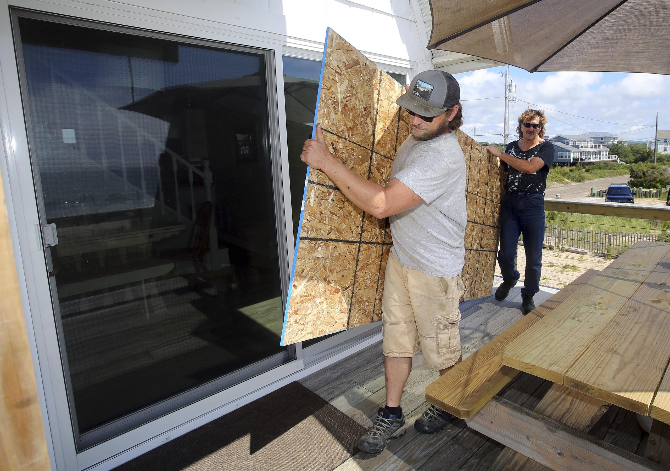 James Masog, al centro, y Gary Tavares, llevan una tabla de conglomerado para proteger las puertas de vidrio de la casa de un cliente en Charlestown, Rhode Island, antes de la llegada del huracán Henri.