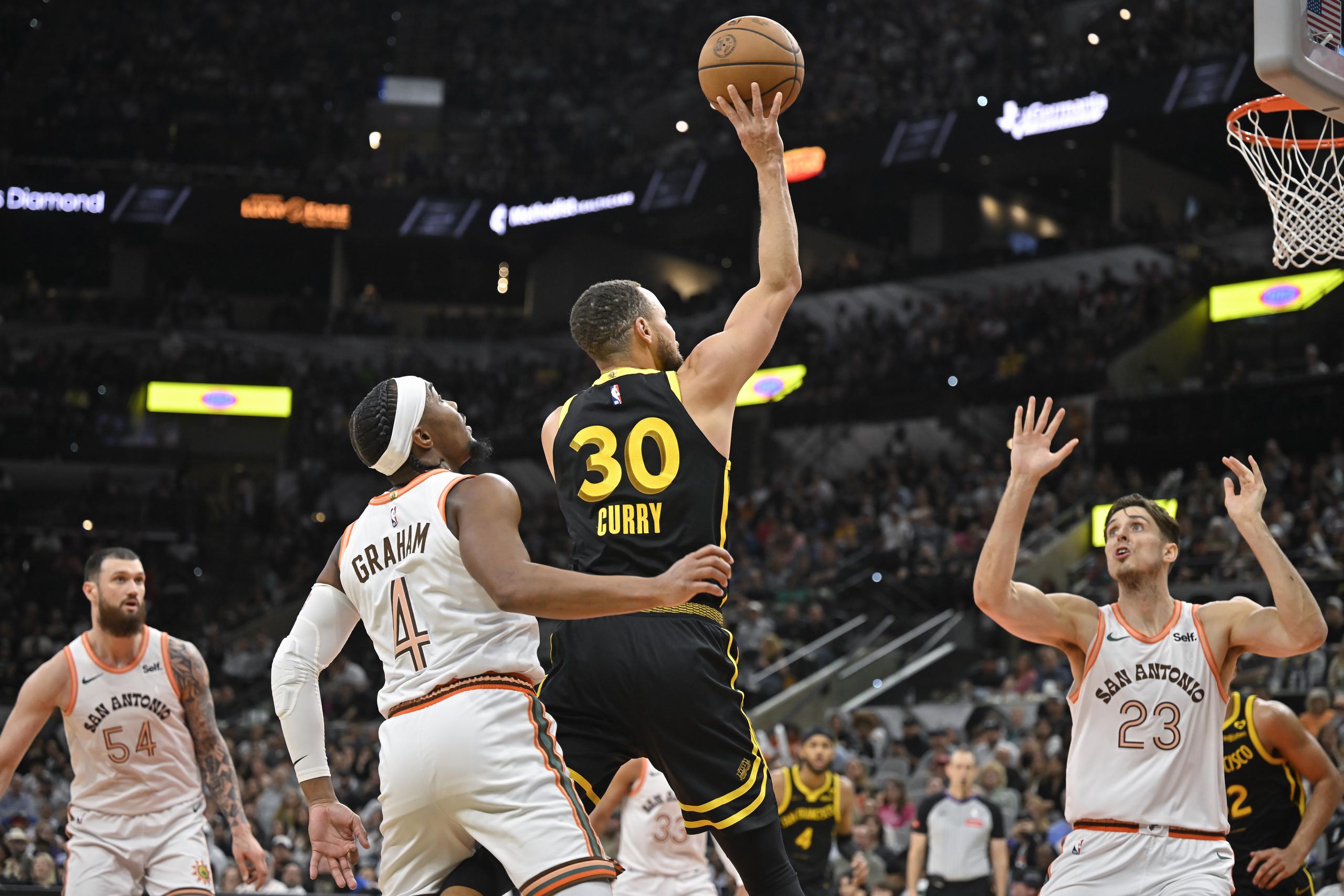 Stephen Curry (30), de los Warriors de Golden State, dispara ante las miradas de Devonte' Graham (4) y Zach Collins (23), de los Spurs de San Antonio, durante la segunda mitad del juego de baloncesto de la NBA, el domingo 31 de marzo de 2024, en San Antonio. (AP Foto/Darren Abate)