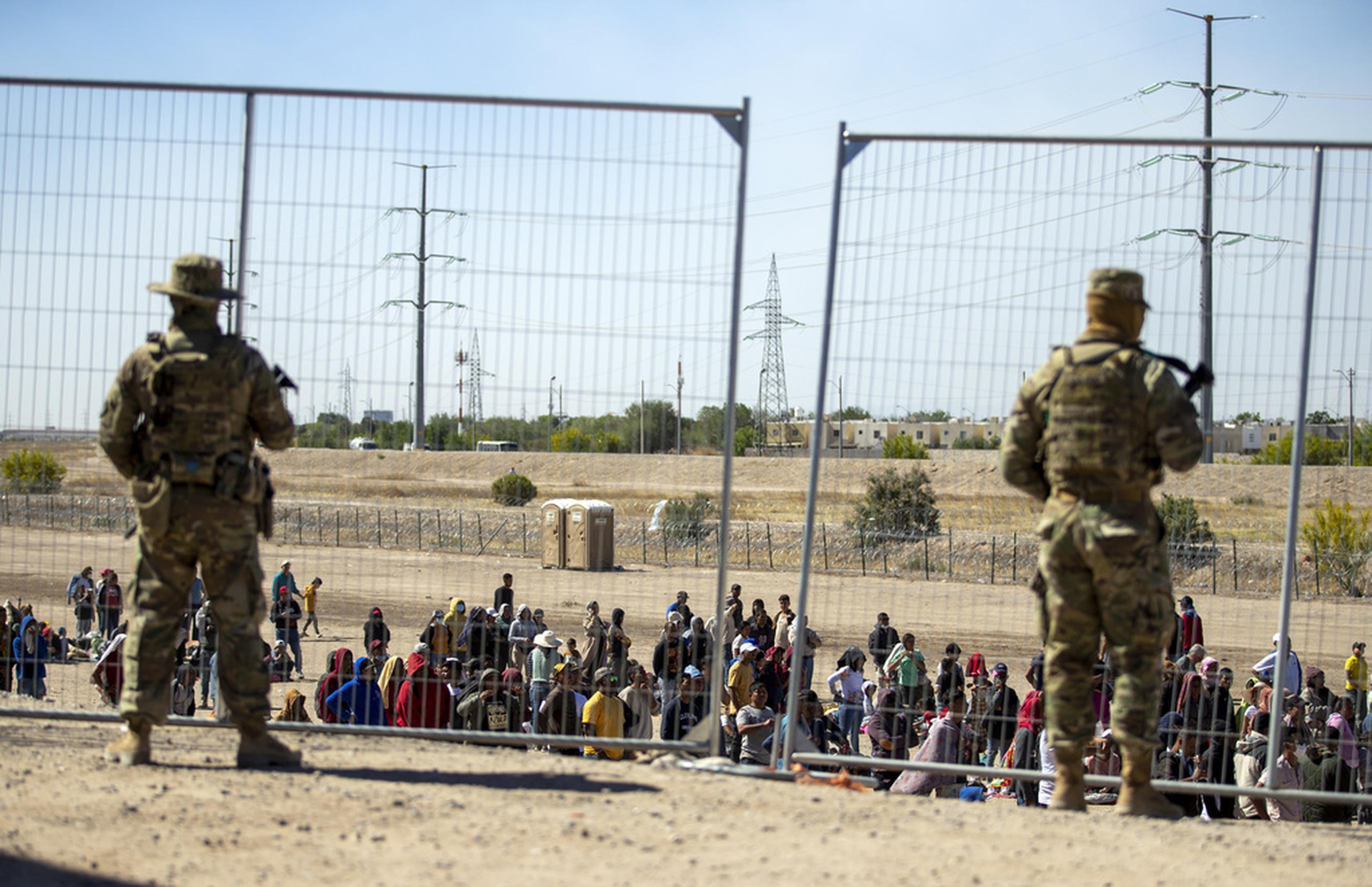 Varios migrantes esperan junto a la cerca fronteriza ante la mirada de guardias nacionales de Texas para entrar en El Paso, Texas, el 10 de mayo de 2023.