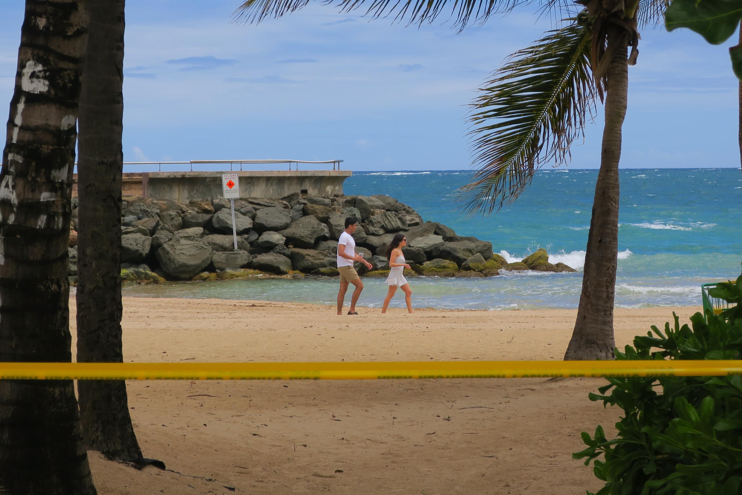 Una pareja camina por la playa pese a la cinta de aviso de "Área restringida" por el sector de Condado en San Juan