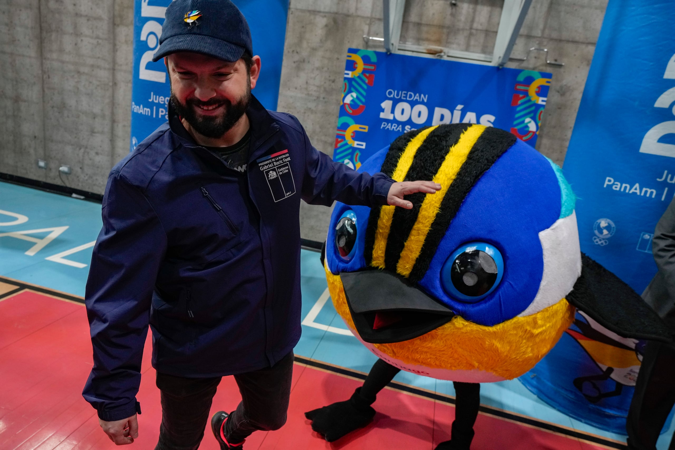 El presidente de Chile Gabriel Boric camina junto a la mascota de los Juegos Panamericanos Fiu, a la llegada al evento que marca los 100 días para el inicio de la justa en el centro deportivo La Pintana en Santiago, Chile el miércoles 12 de julio del 2023. (AP Foto/Esteban Felix)