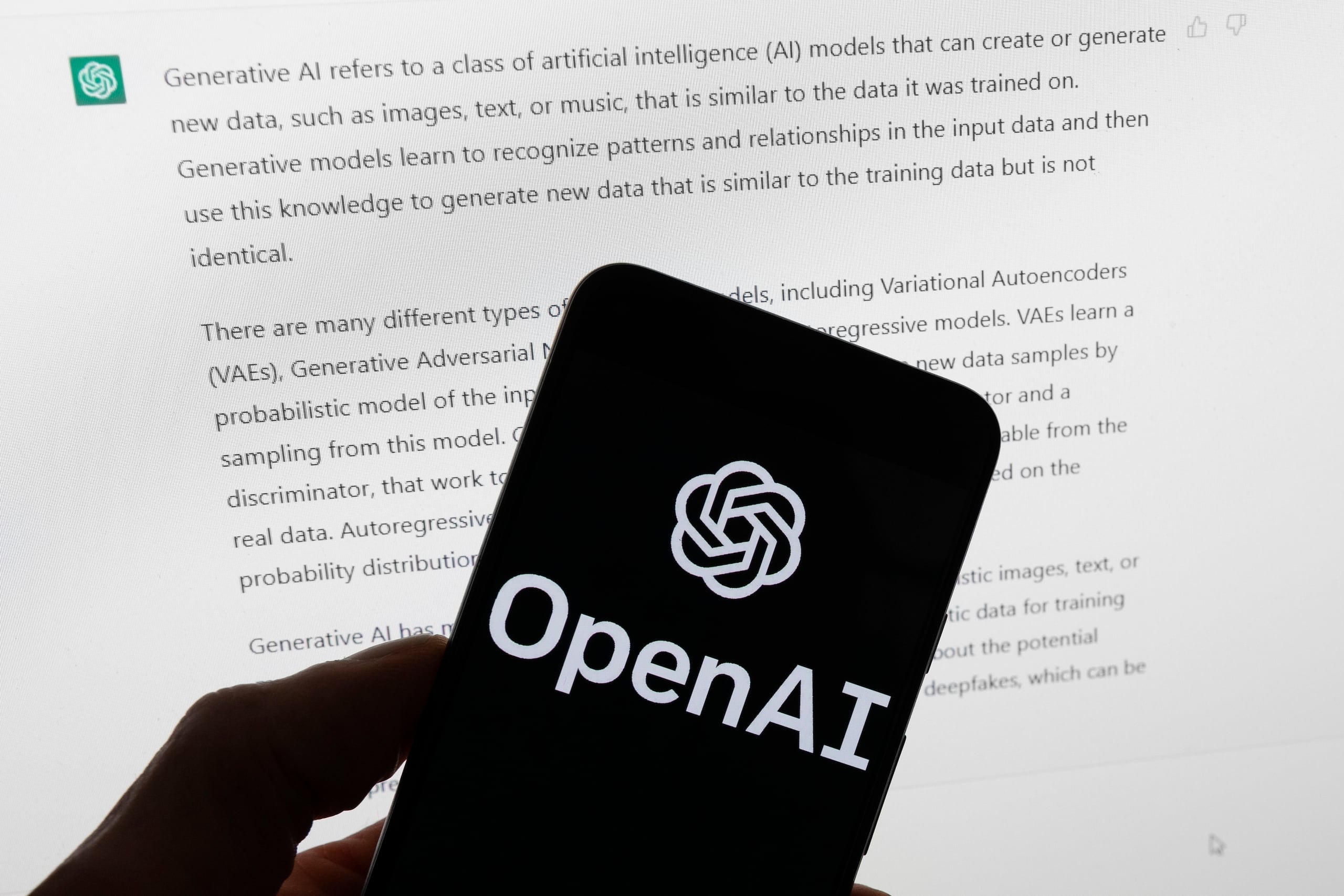 El logotipo de OpenAI se ve en un teléfono celular frente a una pantalla de computadora que muestra un resultado de ChatGPT.