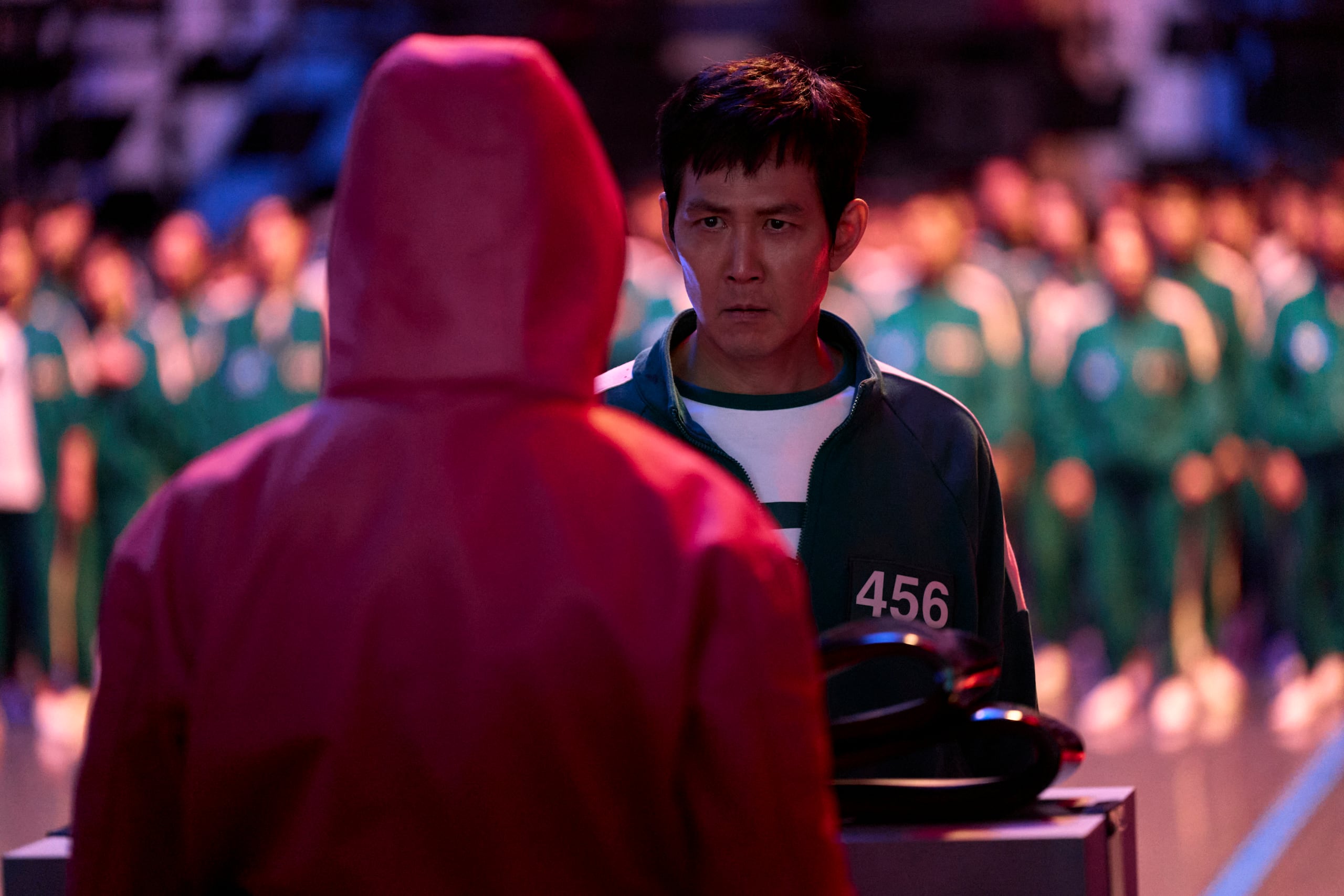 Gi-hun, con su chándal numerado 456, mantiene una conversación tensa con un vigilante en rojo, cuya máscara oculta cualquier emoción. En la primera temporada, desafió las reglas, mostrando su valentía frente al sistema despiadado. Los vigilantes, rápidos en aplicar castigos, controlan a los jugadores con una autoridad implacable. Mientras Gi-hun enfrenta un dilema moral, los participantes en verde esperan, sabiendo que cualquier error puede llevar a la muerte. No Ju-han/Netflix © 2024
