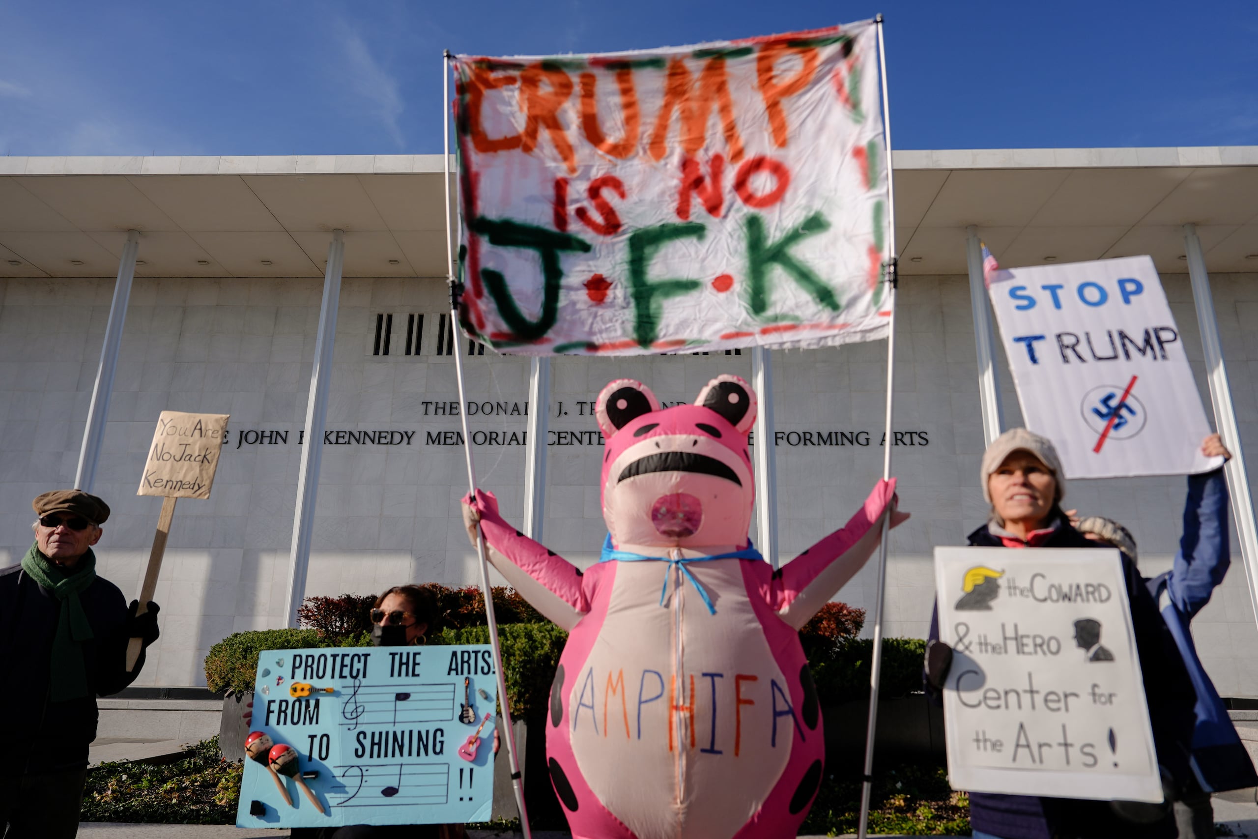 Nadine Siler, de Waldorf Maryland, disfrazada de rana rosa, y otros manifestantes sostienen pancartas durante una protesta frente al Centro John F. Kennedy para las Artes Escénicas, un día después de que la junta designada por el presidente Donald Trump aprobó un cambio de nombre del lugar para añadir el de Trump, el sábado 20 de diciembre de 2025, en Washington. (AP Foto/Julia Demaree Nikhinson)