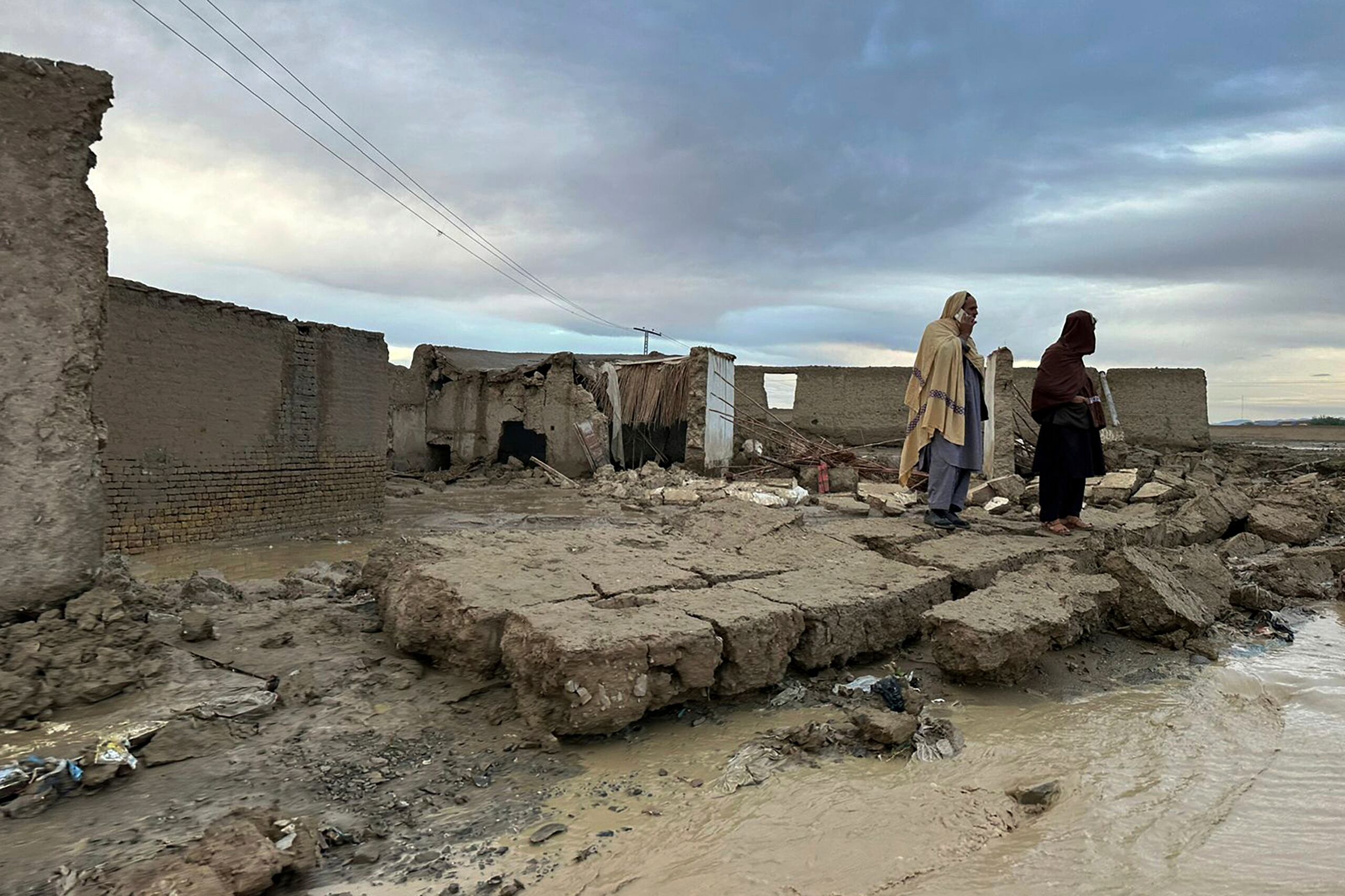 Personas permanecen de pie sobre un muro caído de su vivienda, que sufrió daños debido a las fuertes lluvias en un área cerca de Chaman, Pakistán, el jueves, 18 de abril de 2024. (AP Foto/Habib Ullah)