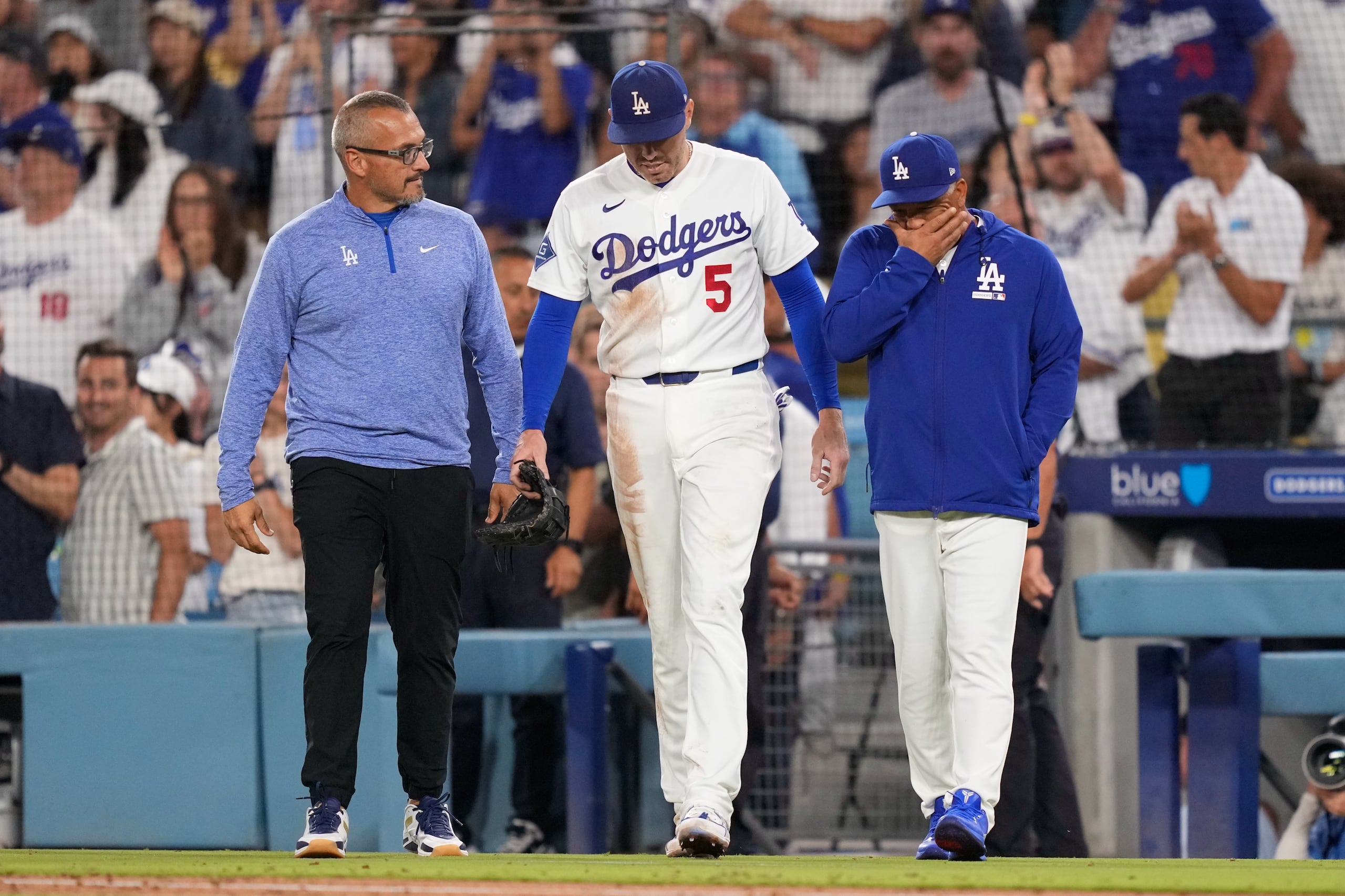 El primera base de los Dodgers de Los Ángeles, Freddie Freeman (al centro), camina de regreso al campo acompañado por el mánager Dave Roberts (a la derecha) y el preparador físico Greg Barajas, después de caer a la cueva al intentar atrapar un elevado de foul conectado por Isaac Collins, de los Reales de Kansas City, durante la octava entrada de un partido de béisbol el miércoles 12 de agosto de 2026, en Los Ángeles. (Foto AP/Mark J. Terrill)