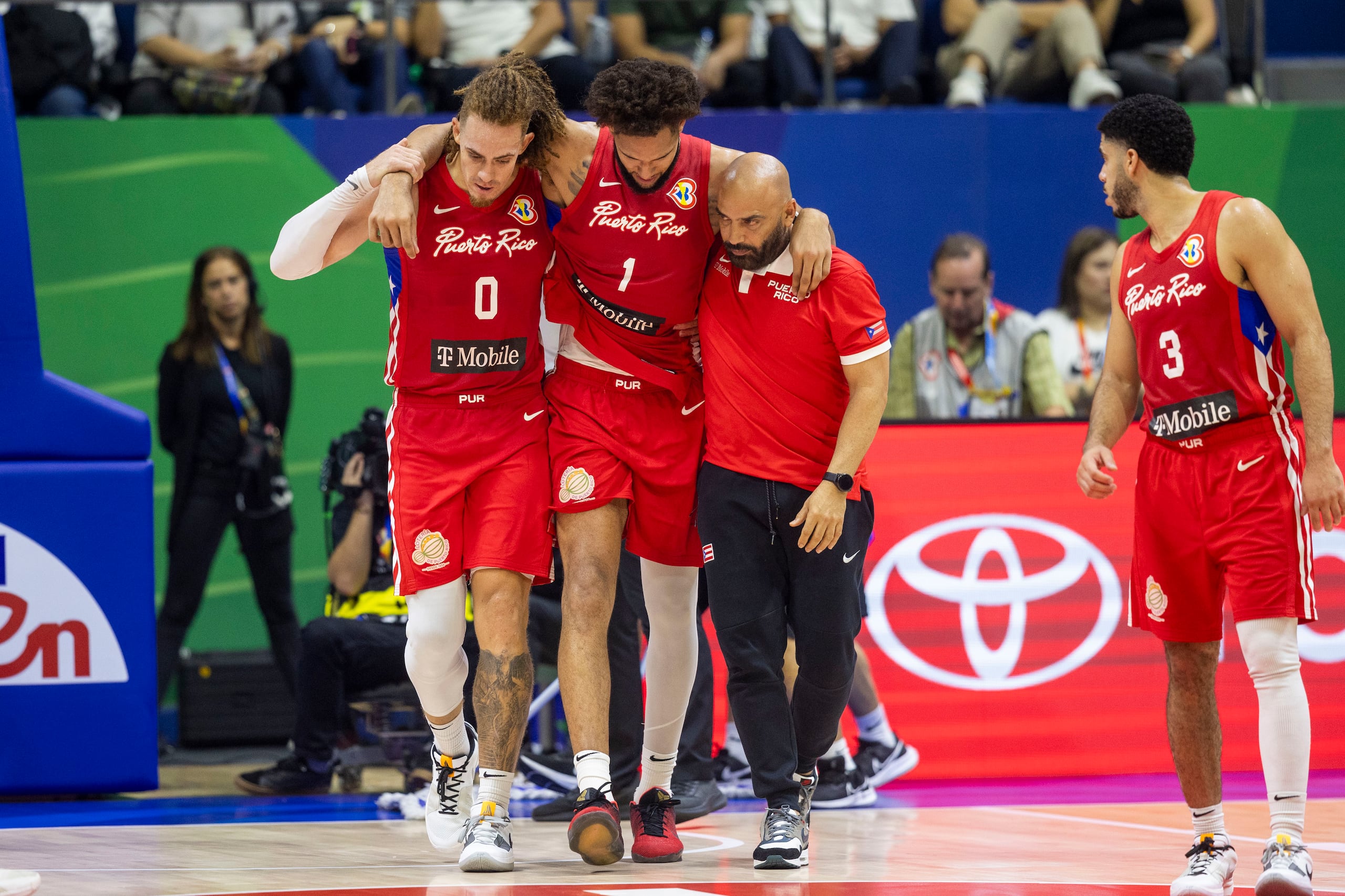 Isaiah Piñeiro y el trainer José Cruz cargan a George Conditt hacia el banco luego de sufrir una torcedura de tobillo hacia la parte final del partido.