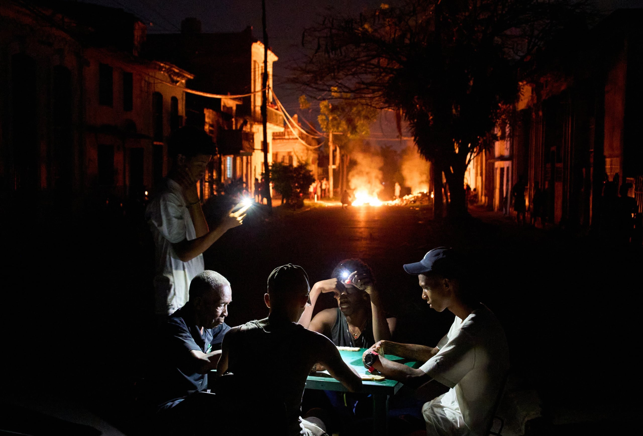 Varias personas se iluminan con sus teléfonos mientras juegan al dominó, al tiempo que arde en una calle de La Habana, Cuba, una hoguera provocada por los vecinos que protestaban contra los prolongados cortes de luz. (AP Foto/Ramón Espinosa)