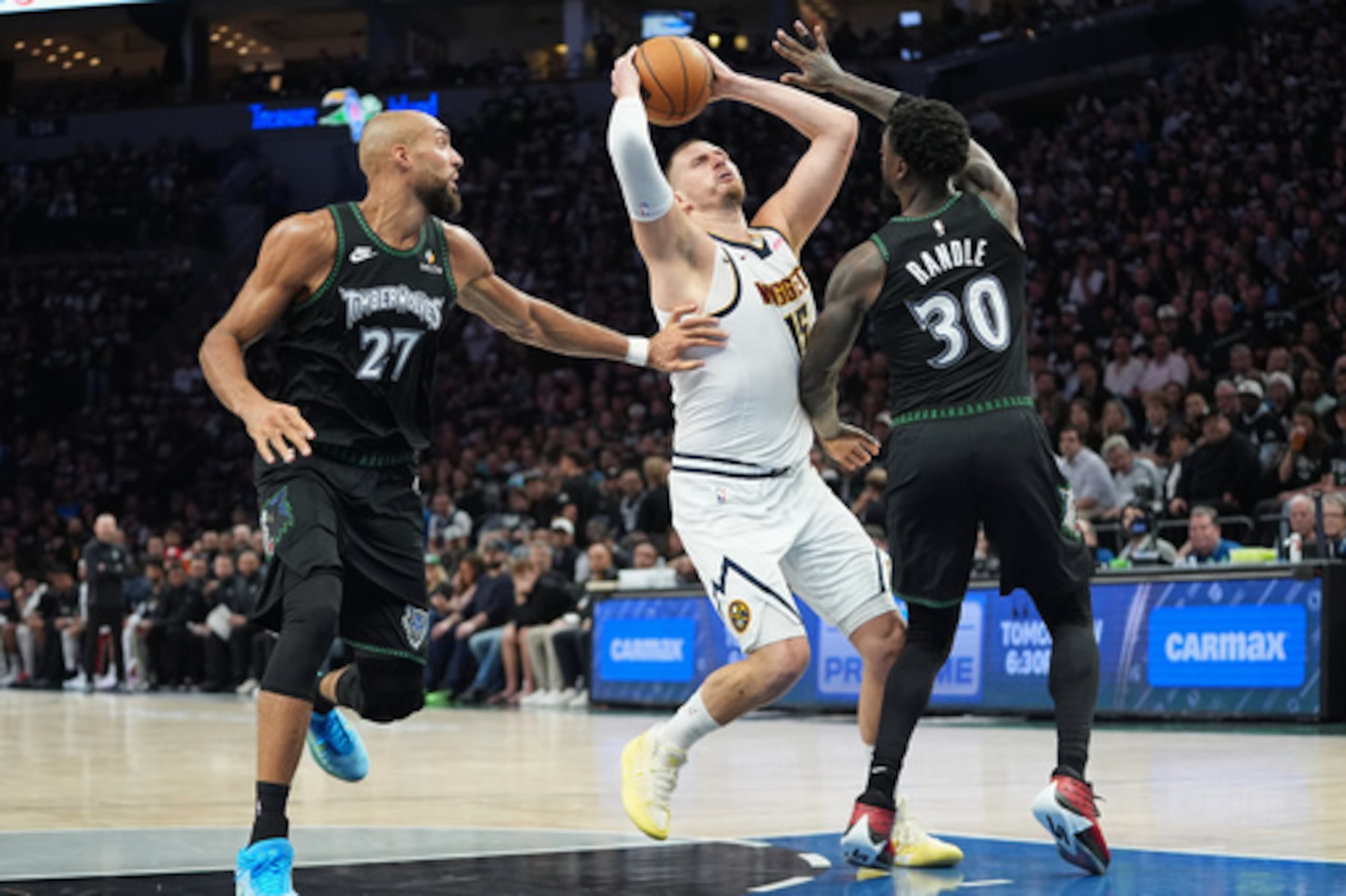 El pívot de los Denver Nuggets Nikola Jokic (15) trabaja hacia la canasta mientras el pívot de los Minnesota Timberwolves Rudy Gobert (27) y el alero Julius Randle (30) defienden durante la primera mitad del tercer partido de la primera ronda de los playoffs de baloncesto de la NBA, el jueves 23 de abril de 2026, en Minneapolis. (AP Photo/Abbie Parr)