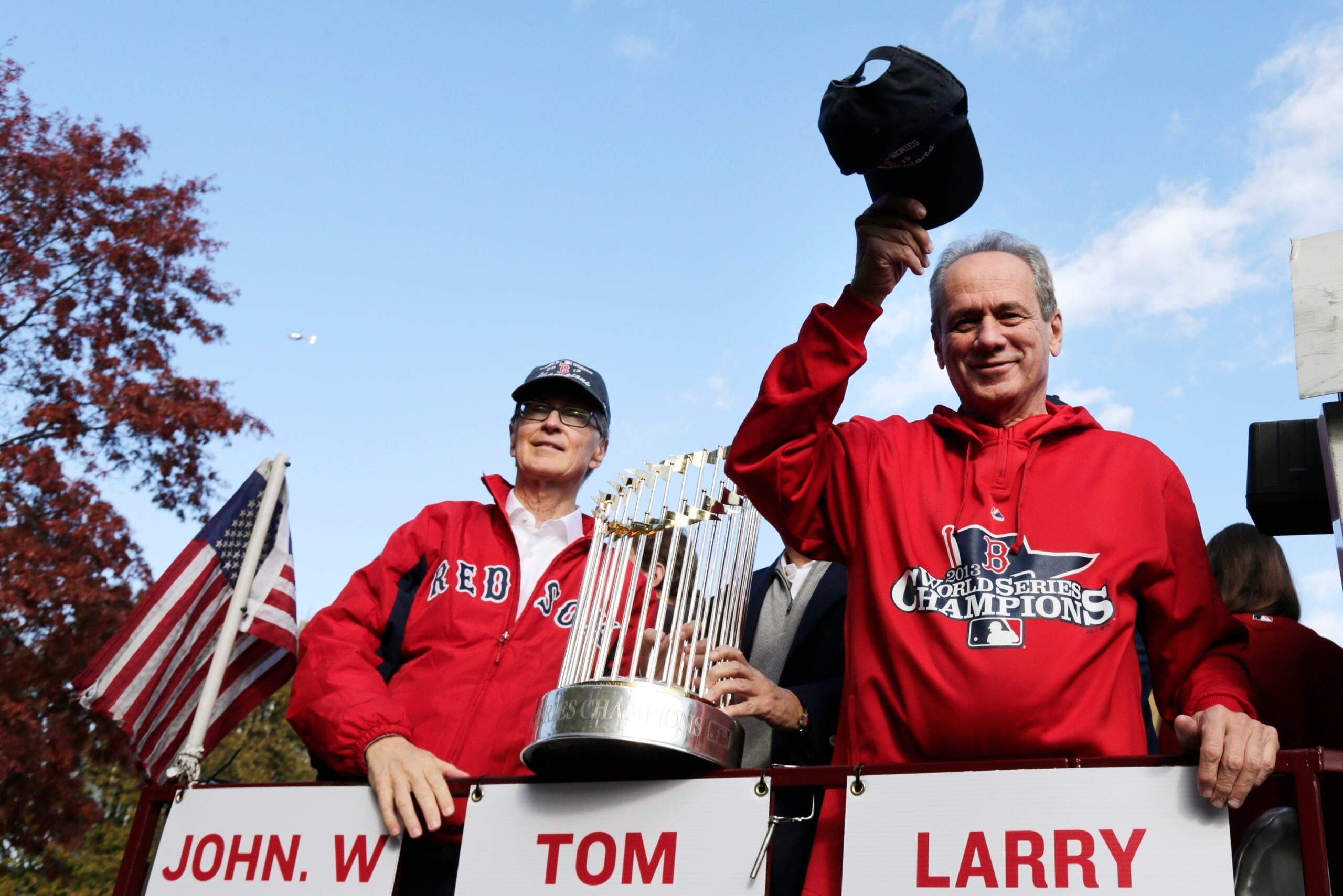 El presidente de los Red Sox de Boston en el 2013 lo era Larry Lucchino, quien aparece a la derecha saludando a los fanáticos junto al dueño John Henry durante el desfile de celebración por la consagración en la Serie Mundial de ese año.
