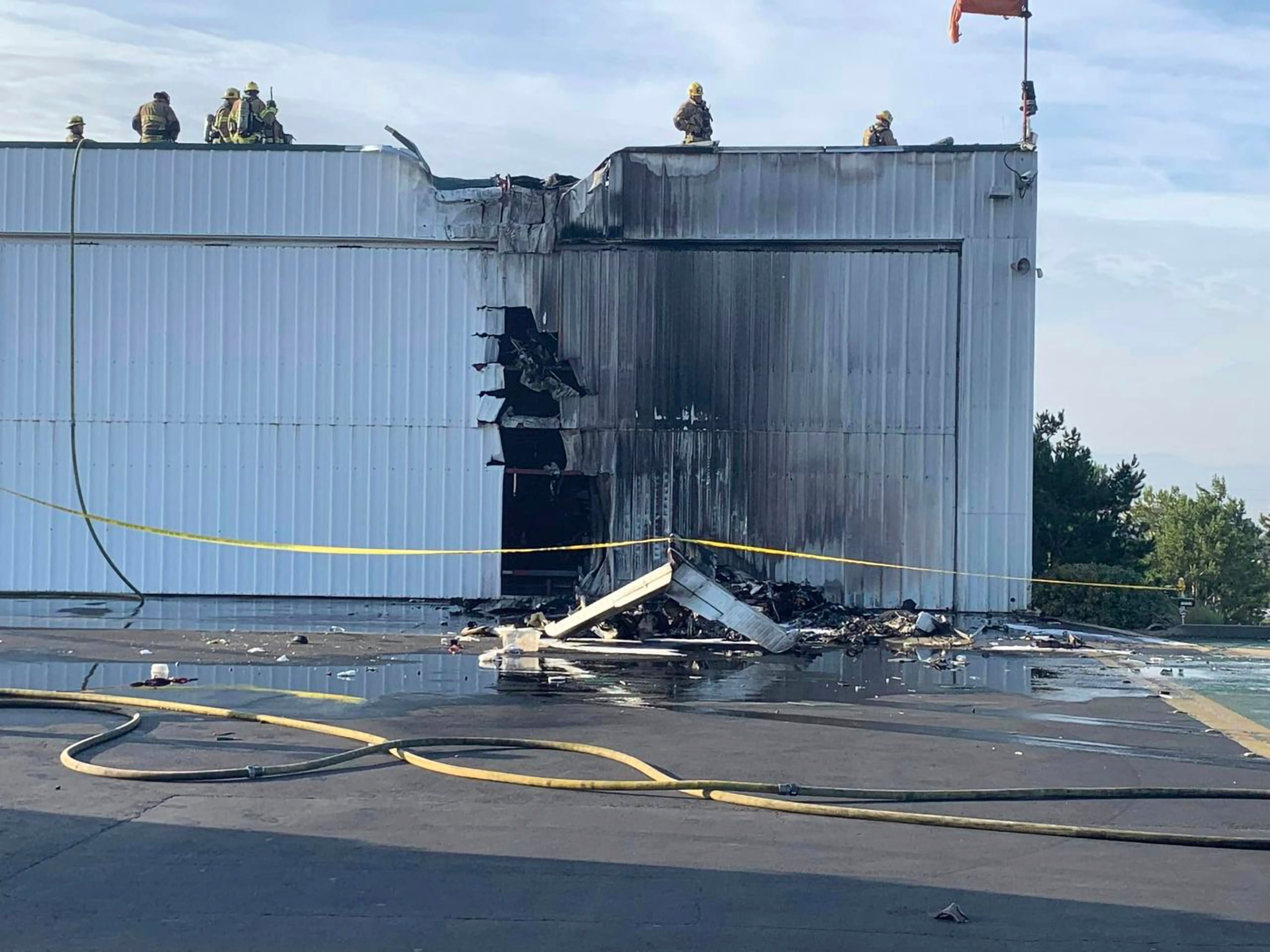 Esta fotografía proporcionada por el Departamento de Bomberos del condado San Bernardino muestra a varios bomberos examinando un hangar dañado por un avión pequeño que se estrelló contra él, el domingo 30 de julio de 2023, en el Aeropuerto Cable en Upland, California. (Departamento de Bomberos del condado San Bernardino vía AP)