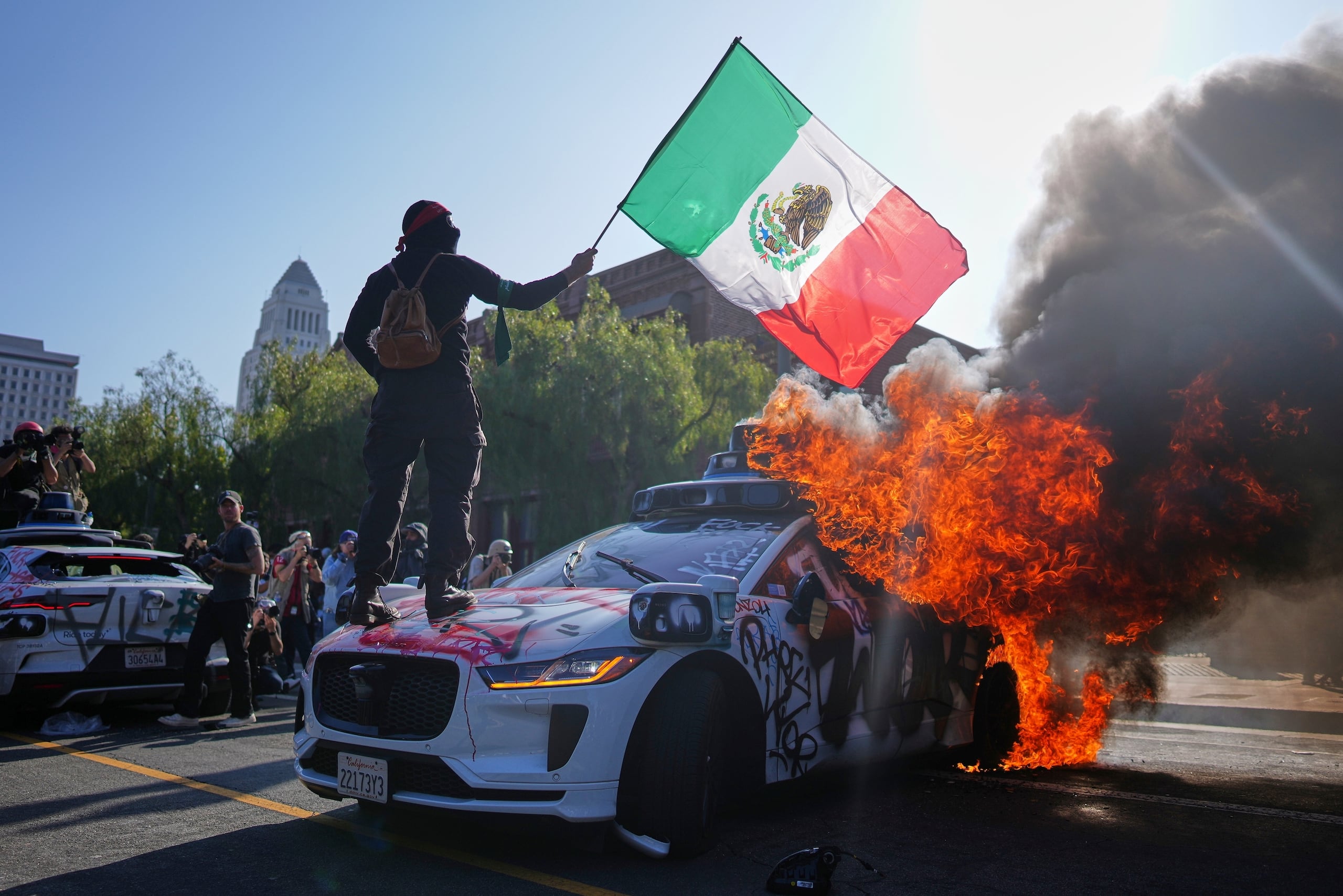 Un manifestante se para sobre un taxi Waymo en llamas cerca del centro de detención metropolitano del centro de Los Ángeles, domingo 8 de junio de 2025, tras la protesta de anoche contra la redada de inmigración.