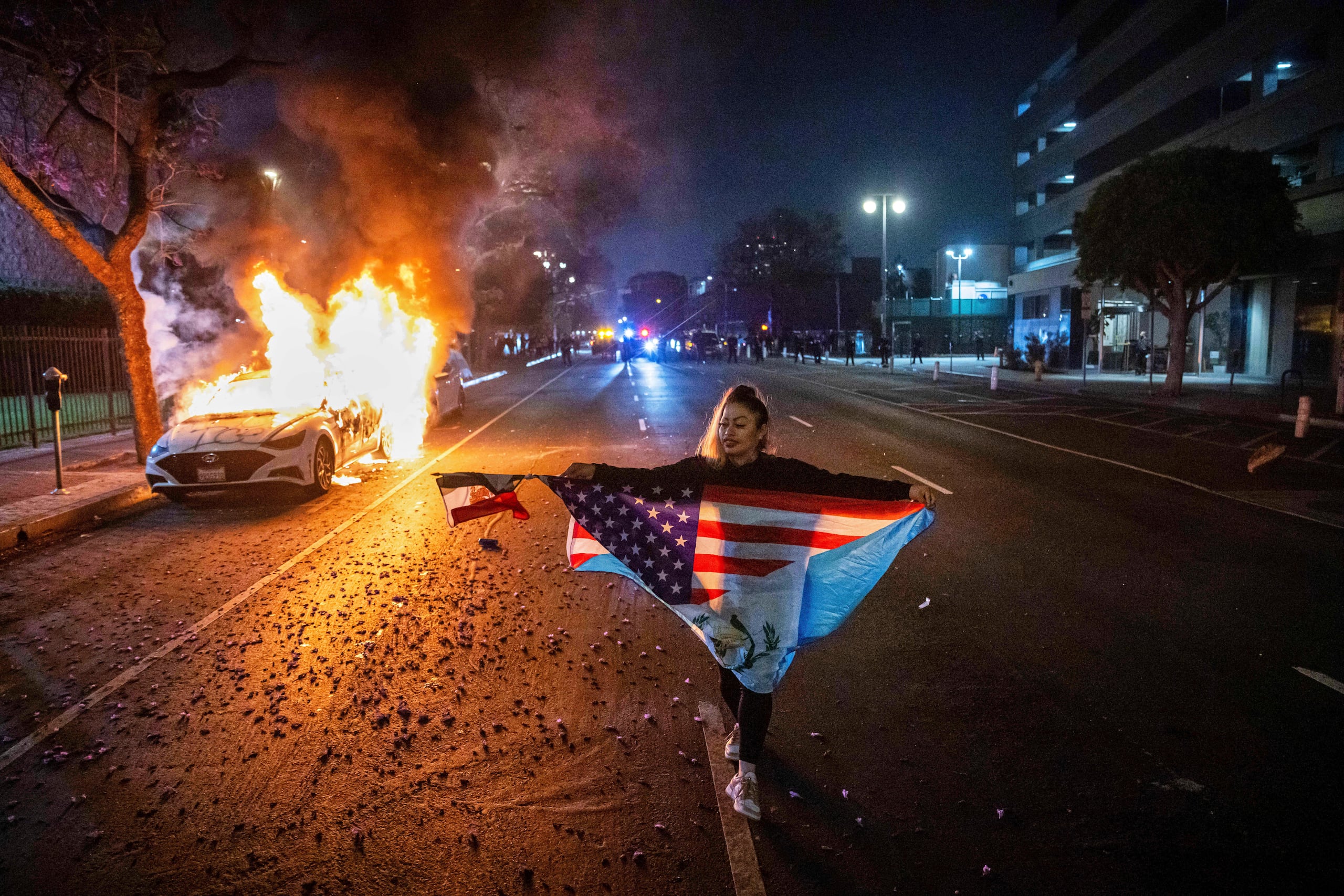 Una persona con varias banderas camina junto a un coche en llamas durante las protestas por las redadas de inmigración de la administración Trump en Los Ángeles, el lunes 9 de junio de 2025.