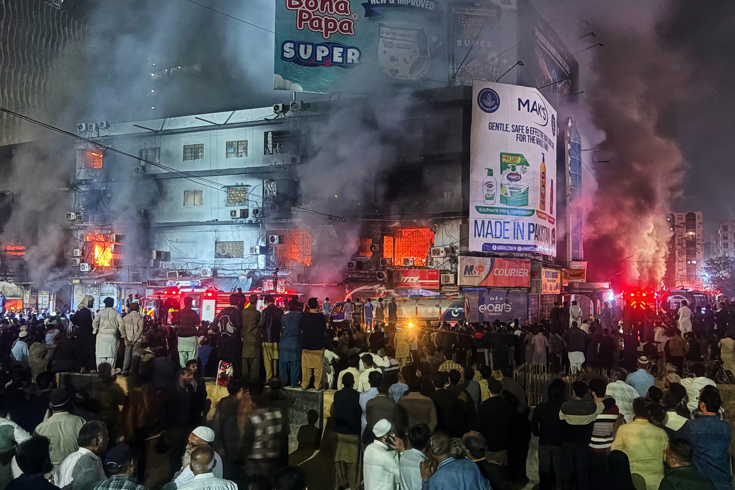 Gente reunida mientras los bomberos intentan conrolar un enorme incendio en un centro comercial de varias plantas en Karachi, Pakistán, el domino 18 de enero de 2026. (AP Foto/Mohammad Farooq)