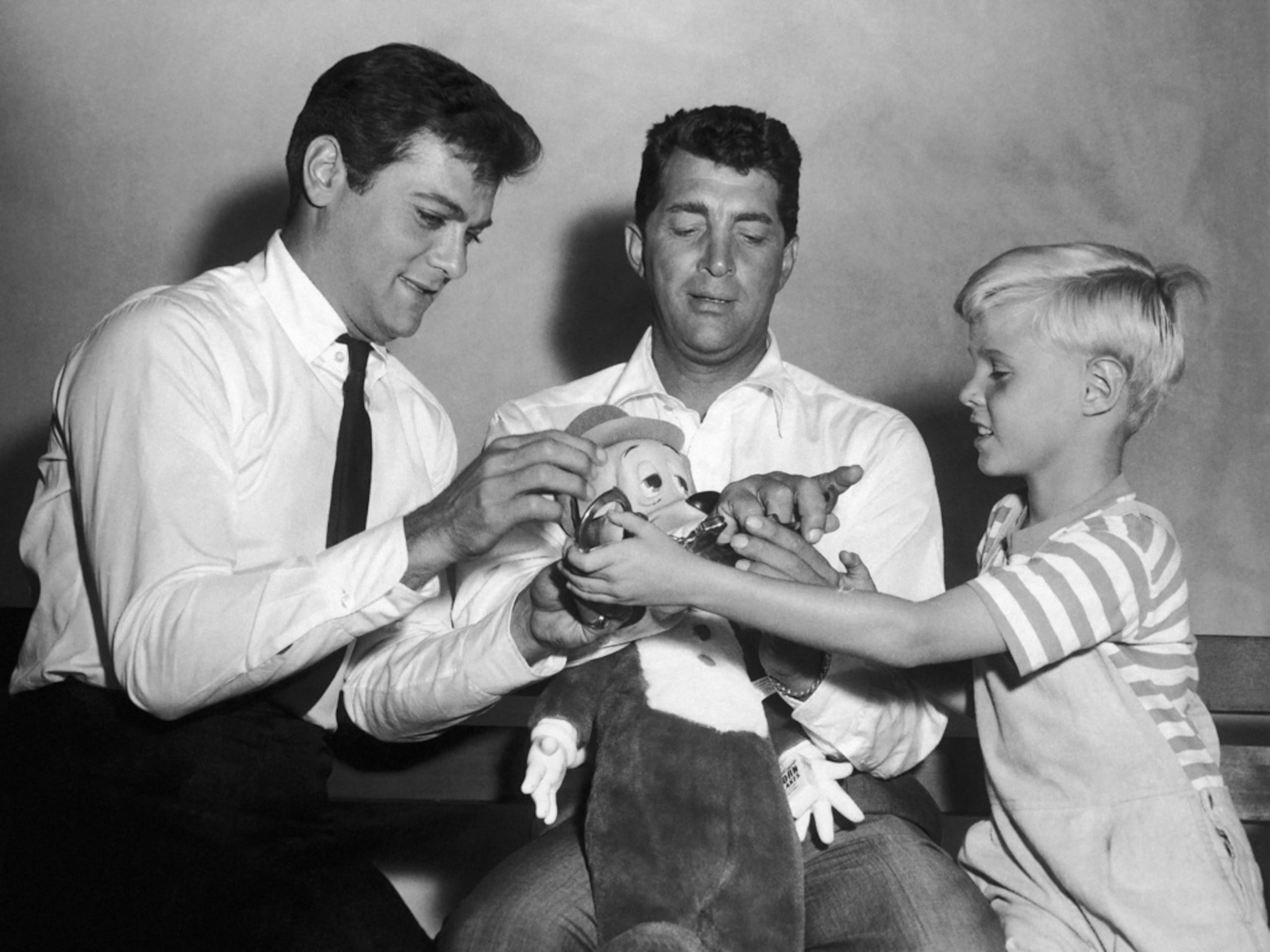 Jay North, joven estrella de la serie "Dennis el travieso", a la derecha, intenta ponerle las esposas a Dean Martin, al centro, mientras juegan con un muñeco de peluche junto a Tony Curtis el 15 de septiembre de 1959.