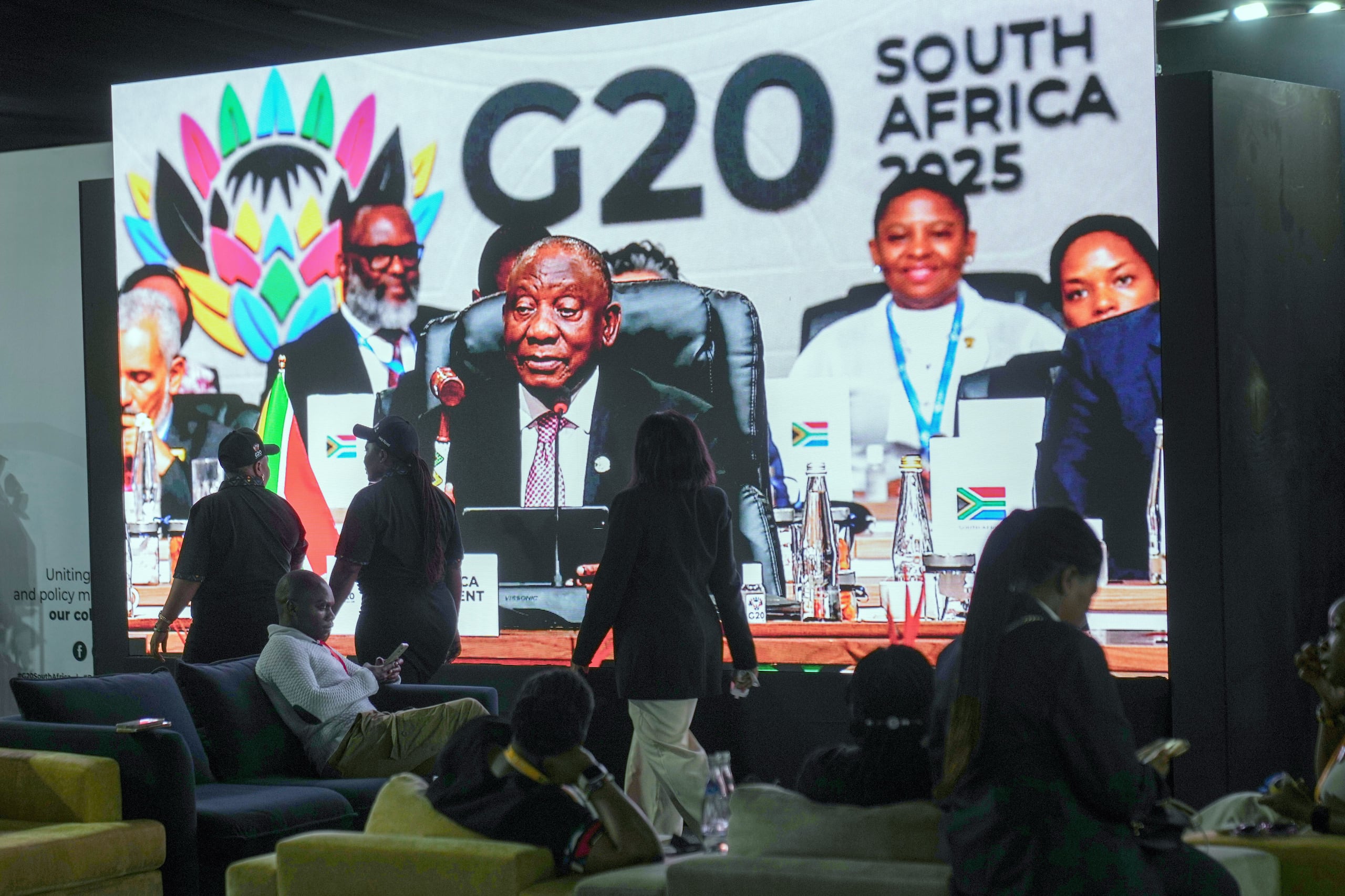 Gente pasa frente a un televisor gigante donde el presidente sudafricano Cyril Ramaphosa sostiene un mazo de madera cuando clausura oficialmente la cumbre de líderes del G20, en Johannesburgo, Sudáfrica, el domingo 23 de noviembre de 2025. (Foto AP/Jerome Delay)