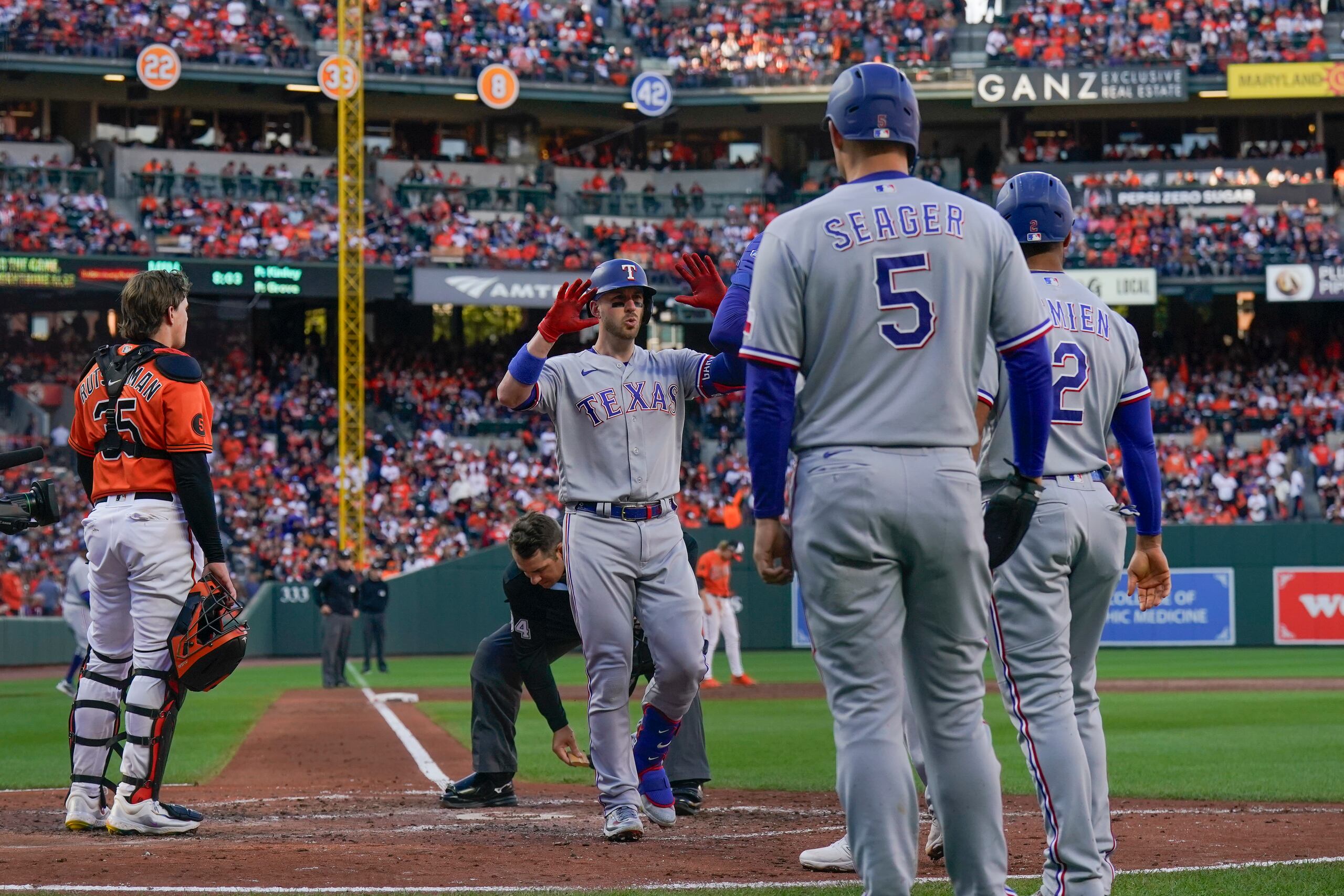 Adley Rutschman, izquierda, receptor de los Orioles de Baltimore, observa como Mitch Garver, centro, de los Rangers de Texas, celebra mientras cruza el plato después de batear un durante la tercera entrada del Juego 2 de la Serie Divisional de la Liga Americana, el domingo 8 de octubre de 2023, en Baltimore. (AP Foto/Julio Cortez)