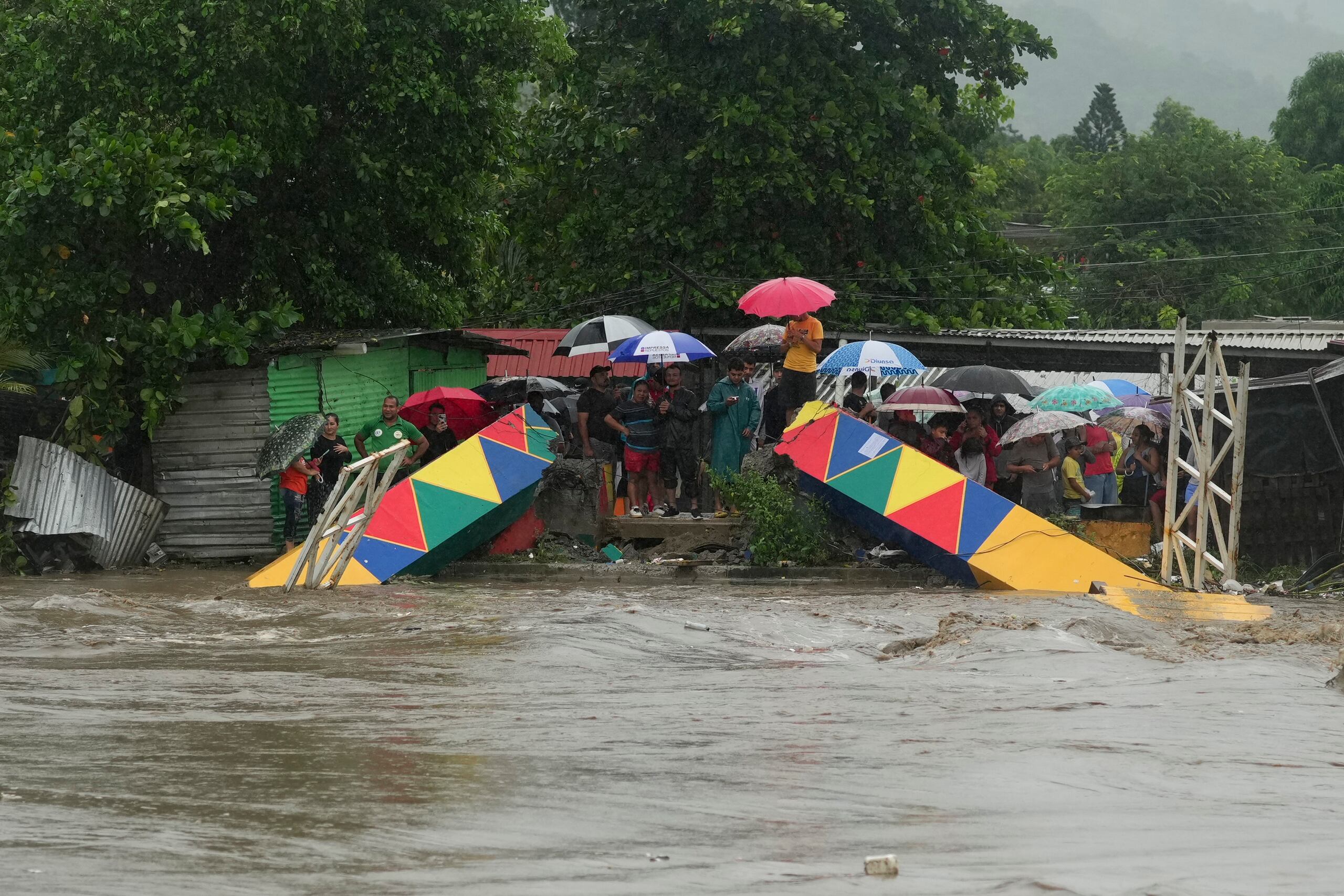 Varias personas paradas frente a un puente peatonal colapsado el sábado 16 de noviembre de 2024 debido a las inundaciones causadas por las lluvias que ha provocado el paso de la tormenta tropical Sara en San Pedro Sula, Honduras. (AP Foto/Moisés Castillo)