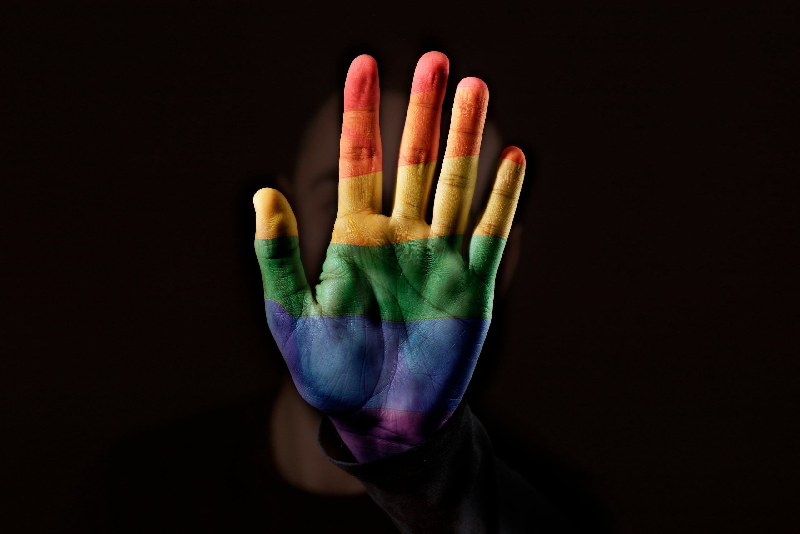 closeup of a young caucasian person with the palm of his or her hand, patterned with a rainbow flag, in front of his or her face, against a black background