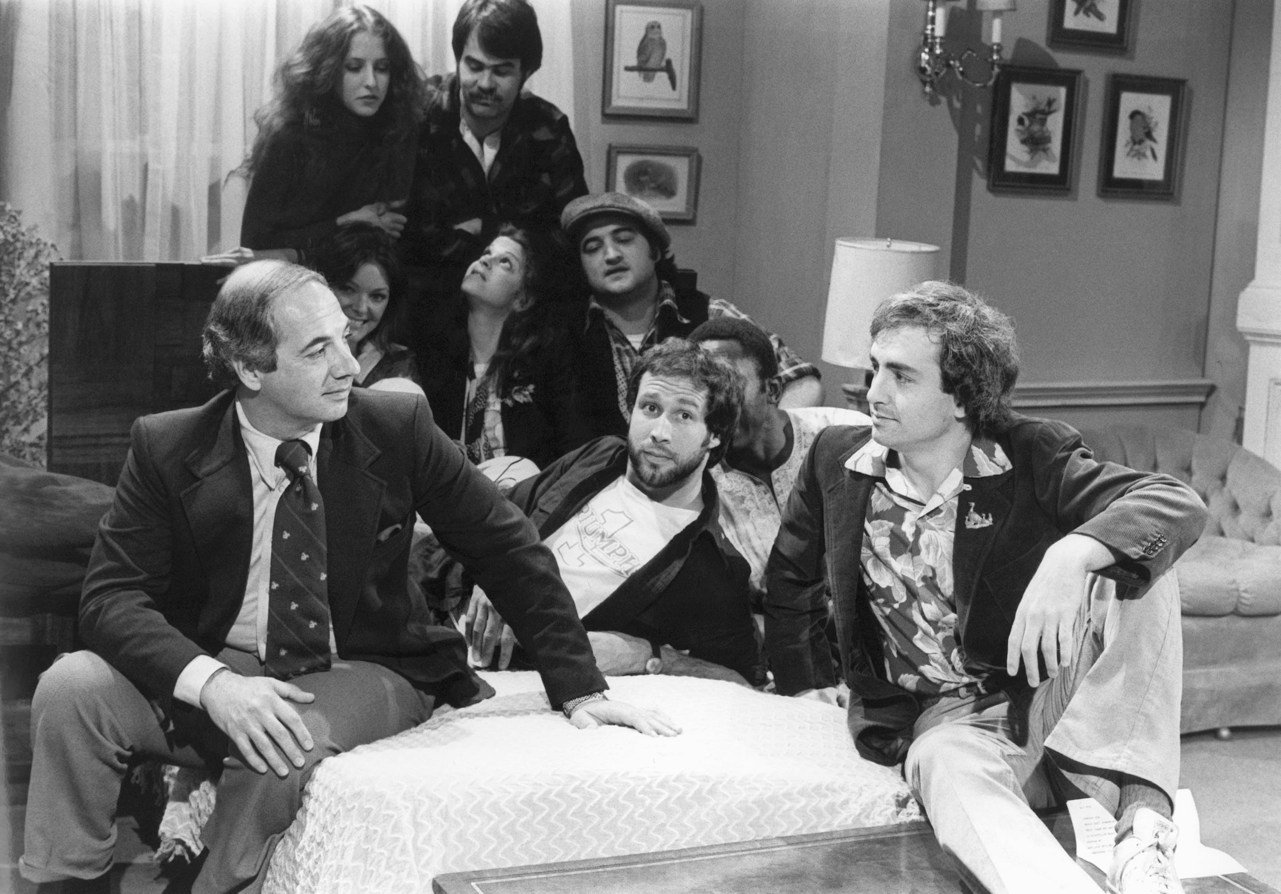 El secretario de prensa de la Casa Blanca, Ron Nessen, a la izquierda, aparece en el set de "Saturday Night Live" con el productor Lorne Michaels, a la derecha, y los miembros del elenco Chevy Chase, en primer plano al centro, Laraine Newman, al fondo a la izquierda, Dan Aykroyd, al fondo a la derecha, Jane Curtain, segunda fila de izquierda a derecha, Gilda Radner, John Belushi y Garret Morris, parcialmente visible, en abril.