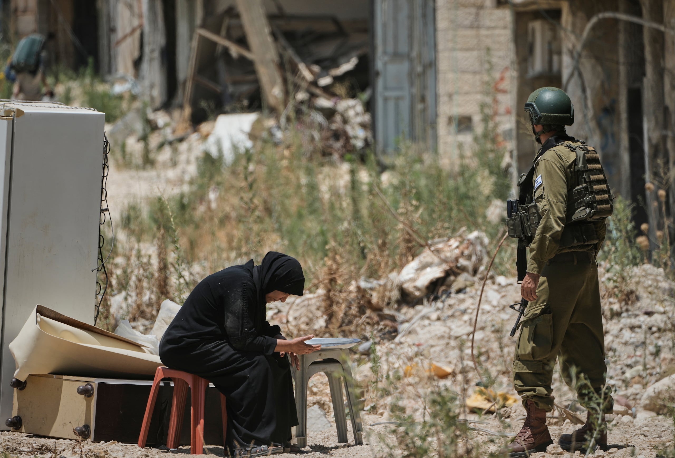 Una mujer descansa sobre un banco, parte de un grupo de palestinos que recogen sus pertenencias de sus hogares evacuados en el campamento de refugiados de Tulkarem, mientras un soldado israelí monta guardia, el miércoles 17 de junio de 2026, en Cisjordania. (AP Foto/Majdi Mohammed)