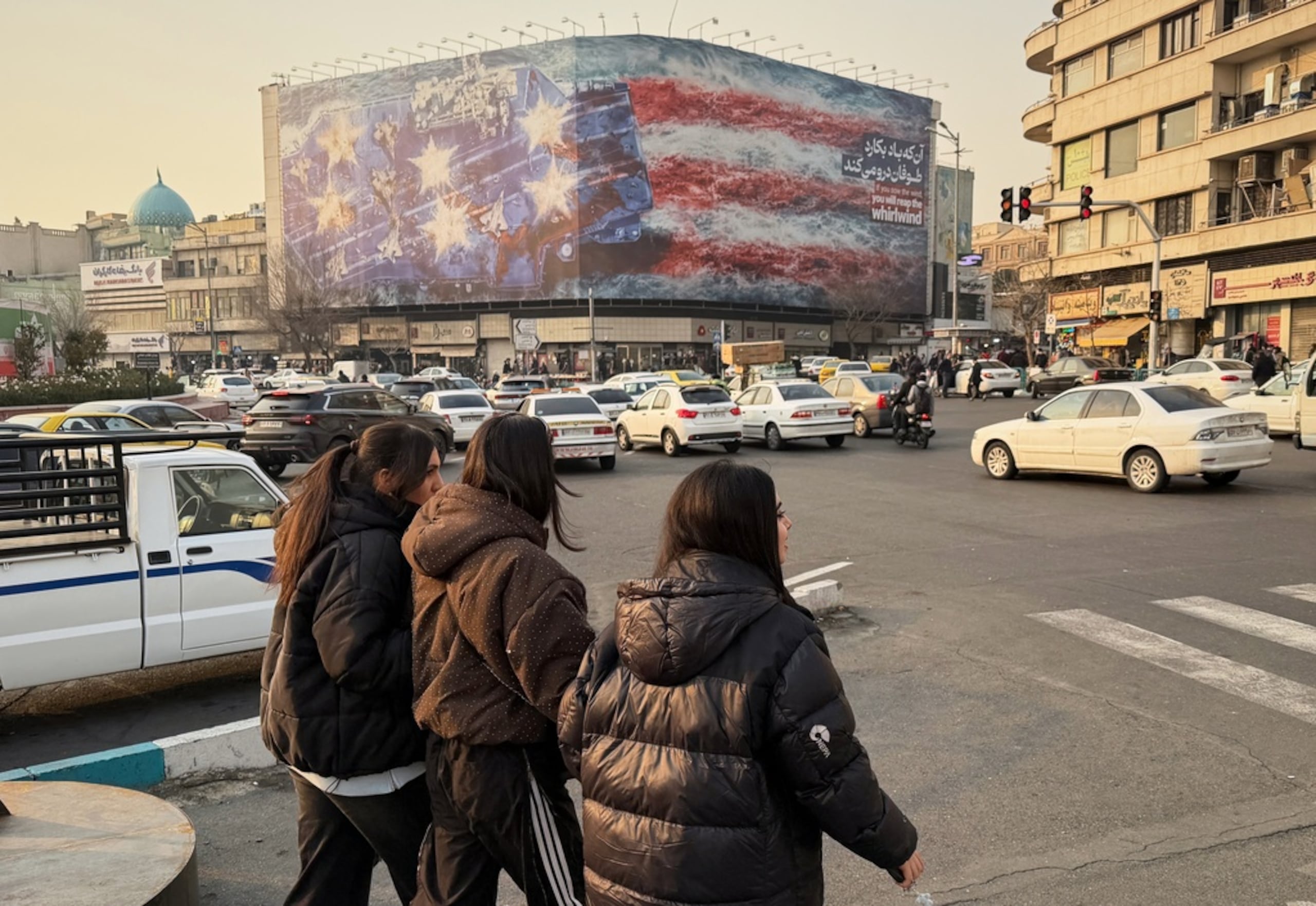 Personas caminan frente a una imagen de un portaaviones estadounidense con aviones dañados en su cubierta y con la leyenda en farsi y en inglés "si siembras viento, cosechas torbellinos", el 25 de enero de 2026, en la Plaza de la Revolución Islámica, en Teherán, Irán.