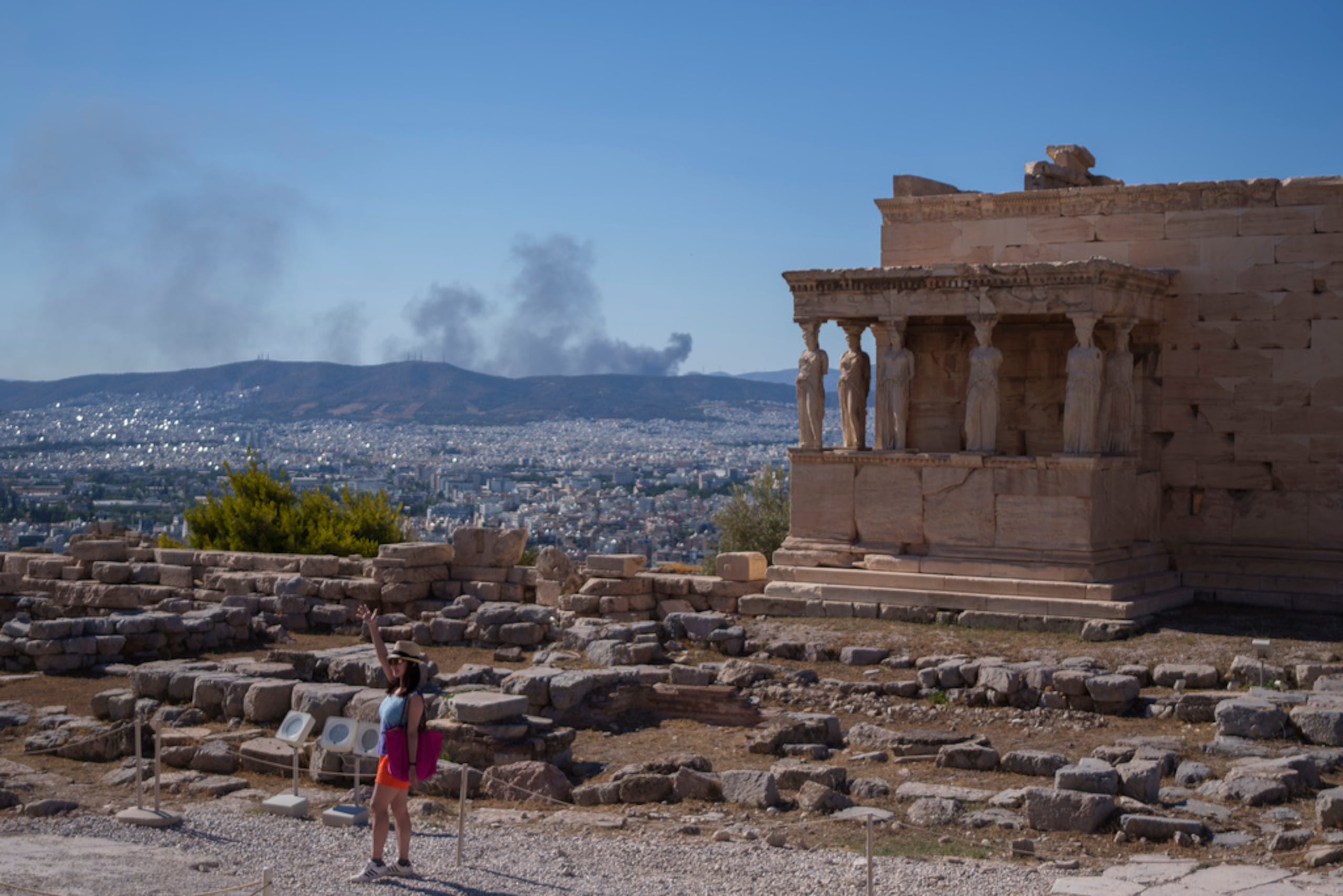 Una mujer posa frente a las estatuas de cariátides del templo Erecteón mientras al fondo se observa humo de un incendio, durante un día caluroso y ventoso en la colina de la Acrópolis, en Atenas, el 16 de julio de 2024.