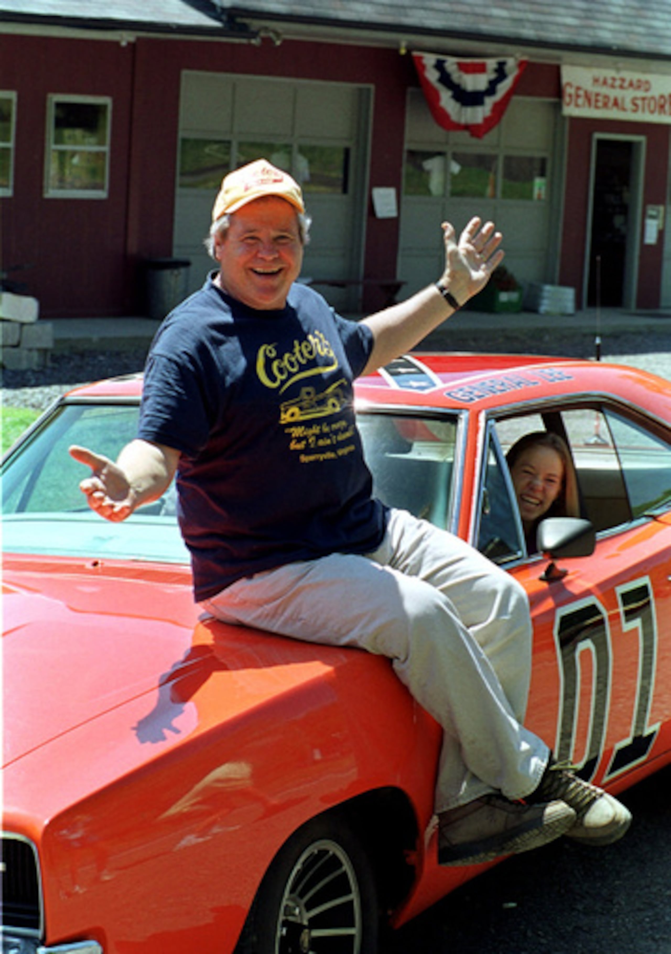ARCHIVO - Cooter, el actor Ben Jones, posa sentado sobre uno de los 229 coches tuneados, llamado «General Lee», utilizados en la serie «Los Dukes de Hazzard», mientras una entusiasta admiradora, Aliceson Johnson, de Charlottesville (Virginia), ocupa el asiento del conductor frente a su tienda en Sperryville (Virginia), el martes 10 de agosto de 1999. (Foto AP/Steve Helber, archivo)