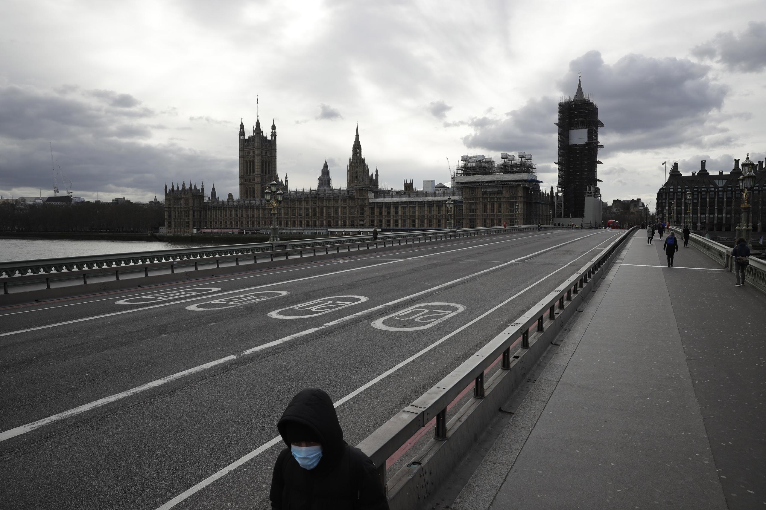 Una persona con una mascarilla en el rostro camina por el puente de Westminster casi vacío con el Parlamento al fondo, en Londres.