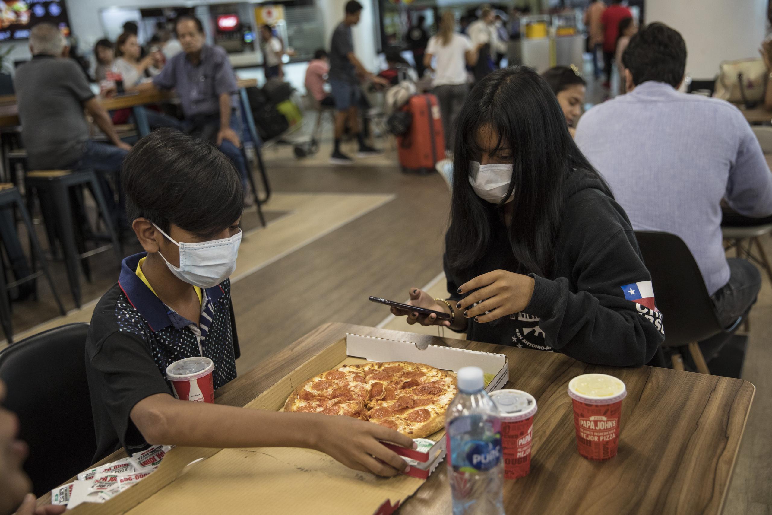 Wearing masks, passenger siblings from Chile, Yhoalibeth Ramos, right, and Oscar Ramos, eat pizza at the international airport in Lima, Peru, Friday, March 6, 2020. Peruvian President Martin Vizcarra announced the first case of the new coronavirus in the country. (AP Photo/Rodrigo Abd)