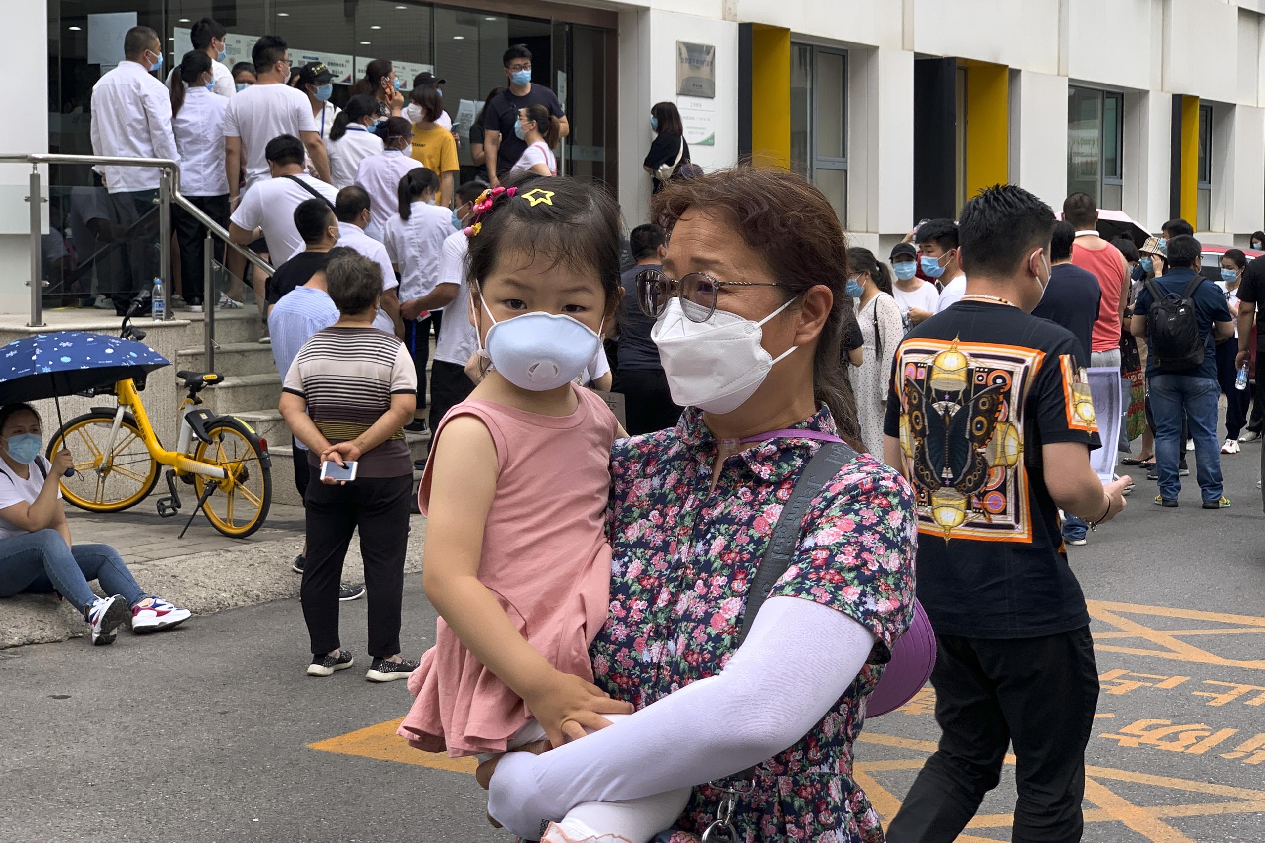 Una mujer carga una niña, ambas con mascarillas, mientras al fondo se obsevra una fila de personas para hacerse las pruebas obligatorias de coronavirus.