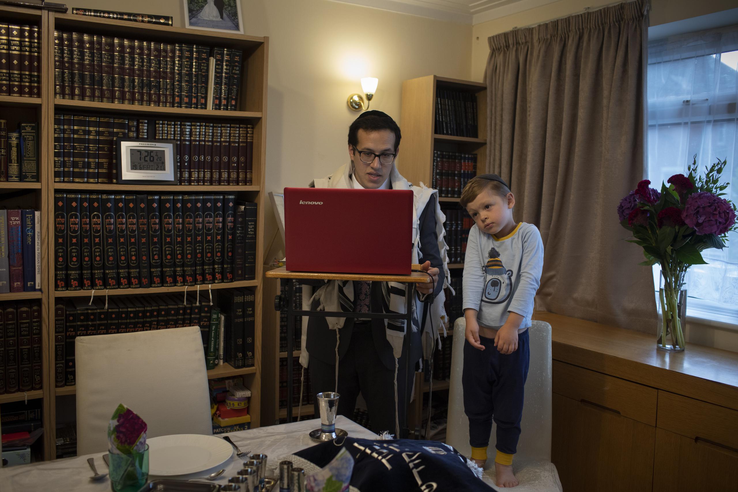 Observado por su hijo Tzvi, de 3 años, el rabino Mordejai Chalk dirige un oficio por teleconferencia desde su casa en Londres